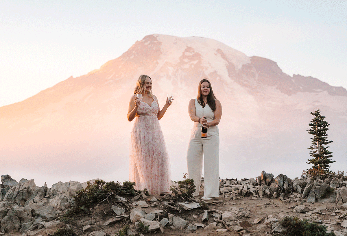 Two women celebrate on a rocky mountain summit with Mt. Rainier glowing pink at sunset behind them.