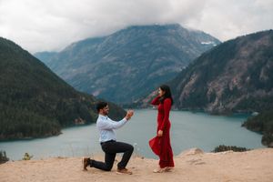 Man proposes to woman in red dress on mountain overlook with scenic lake and misty peaks backdrop.