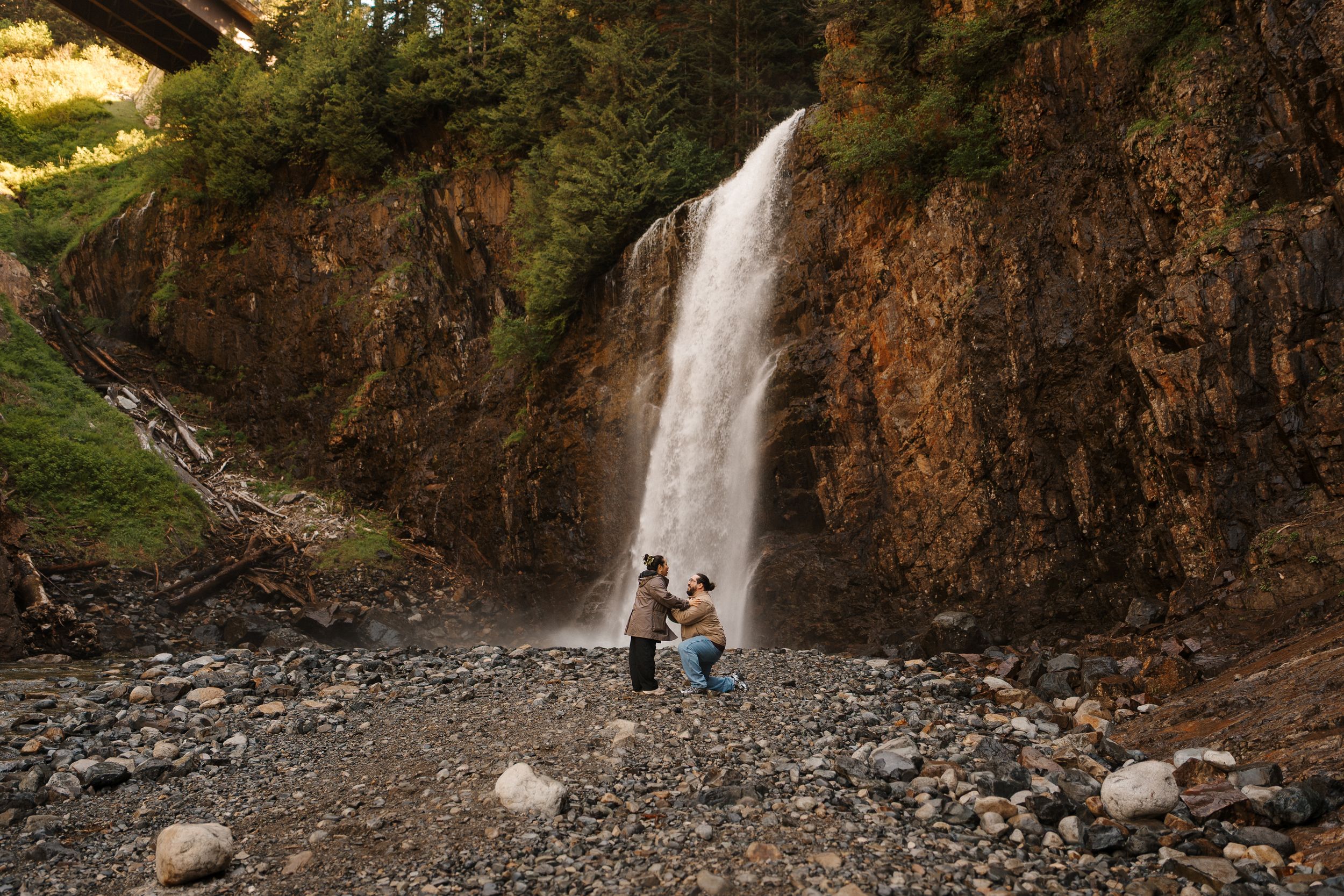 Couple sitting on rocky shore before a tall waterfall surrounded by lush forest and rugged cliffs.