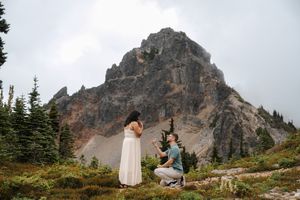 Man proposes to woman in white dress on mountain trail with dramatic rocky peak and evergreen trees in background.