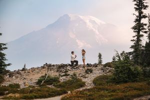 Man proposes to woman on rocky mountain trail with snow-capped Mount Rainier in background at sunset.