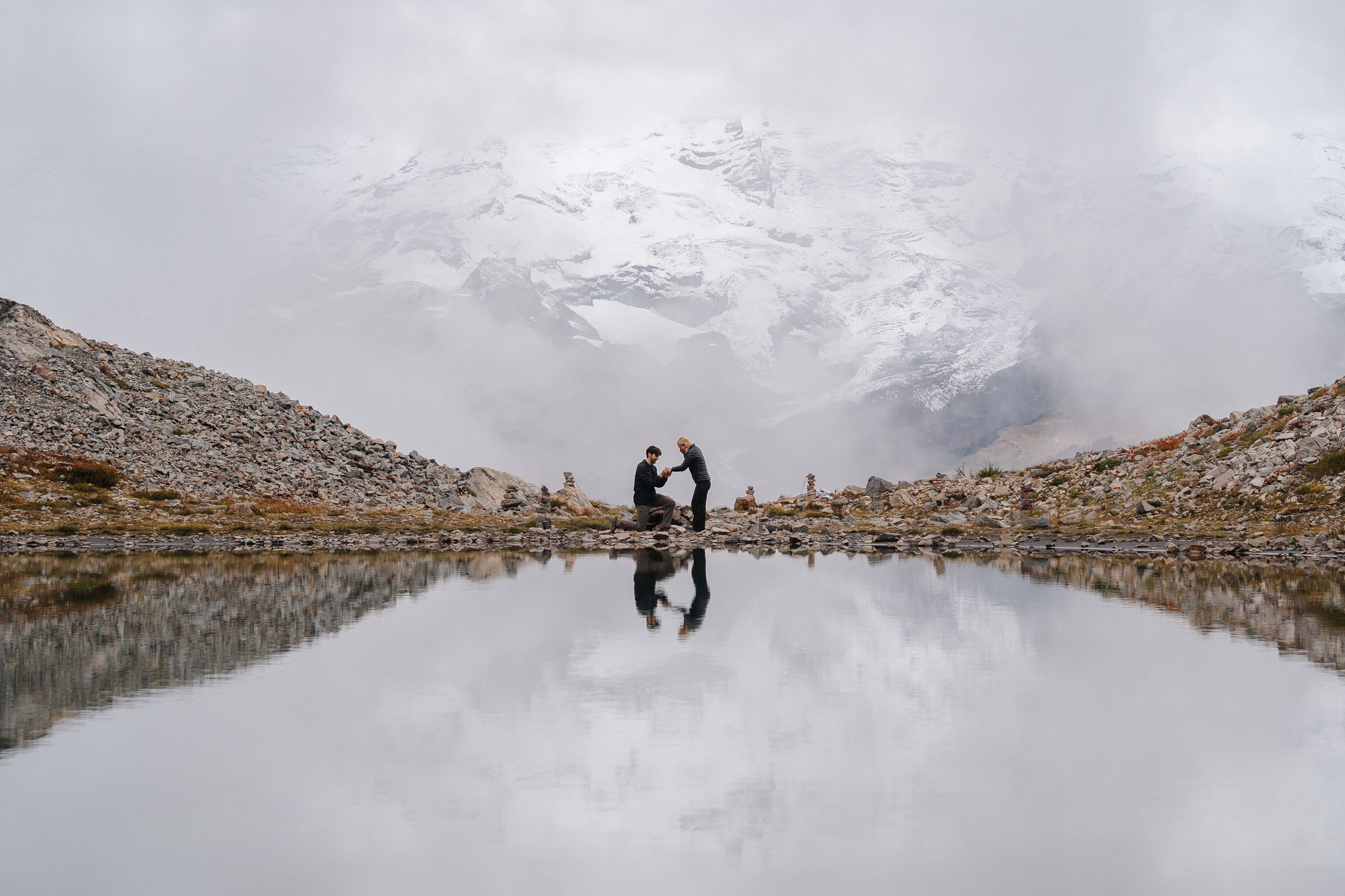 Couple embracing by a reflective alpine lake with snow-capped mountains in the background.