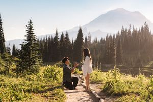 Man proposes to woman on mountain trail surrounded by evergreen trees with scenic peaks in golden sunlight.