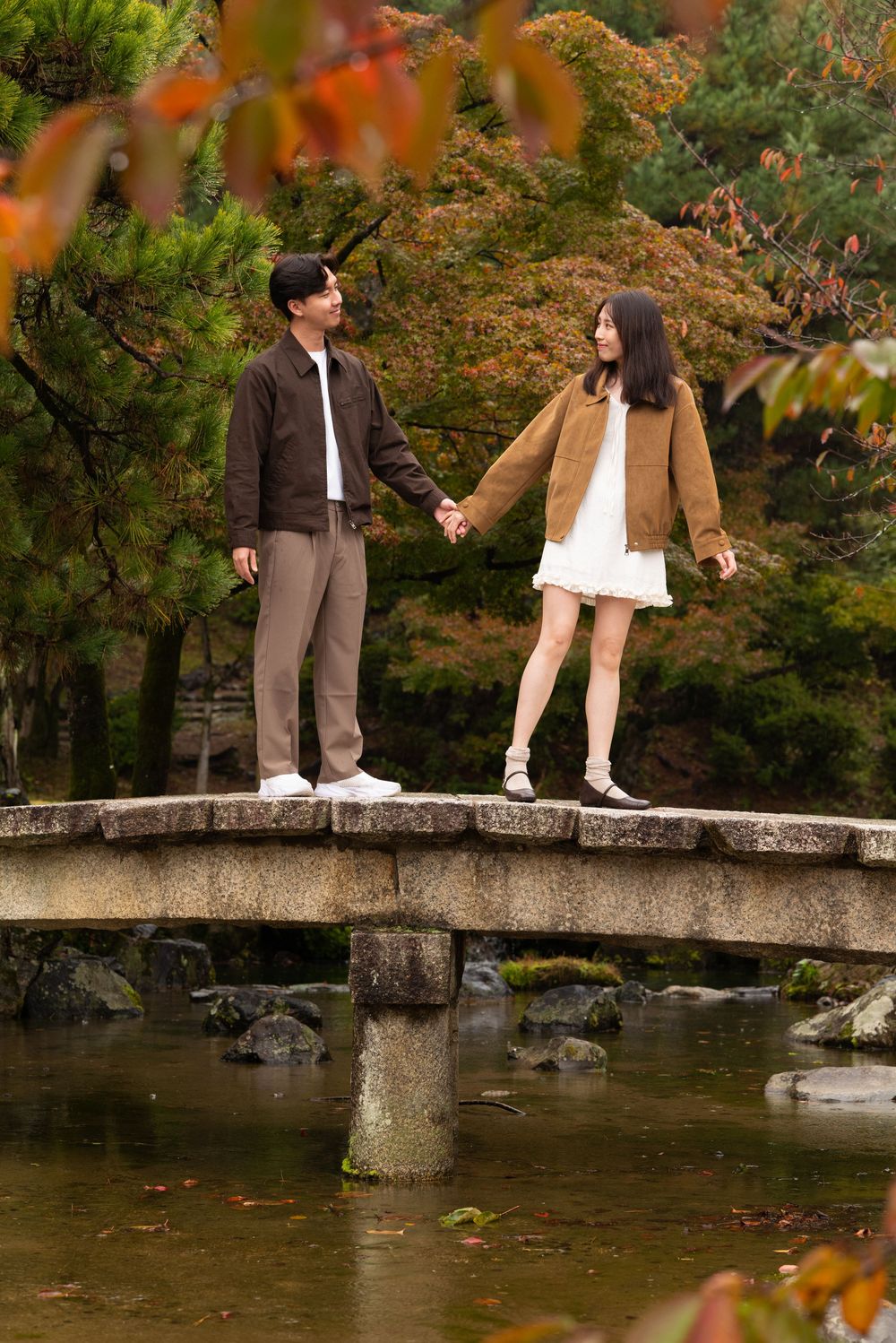 Couple holding hands on a stone bridge surrounded by autumn foliage in a Japanese garden.