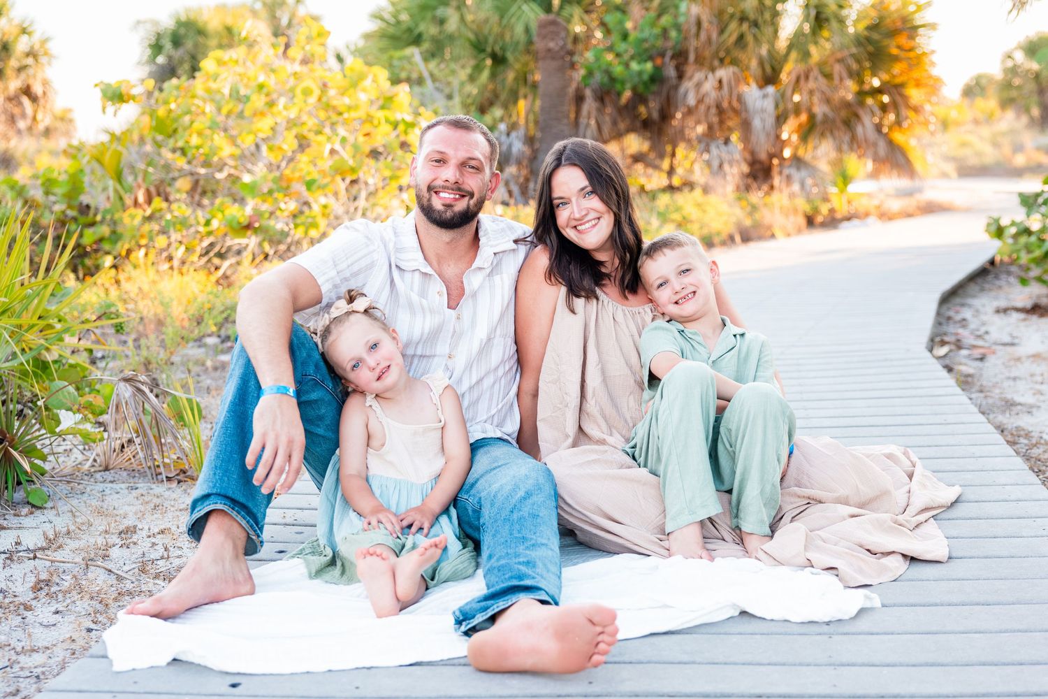 Family of four smiling on a wooden boardwalk surrounded by lush greenery at a sunny beach location.