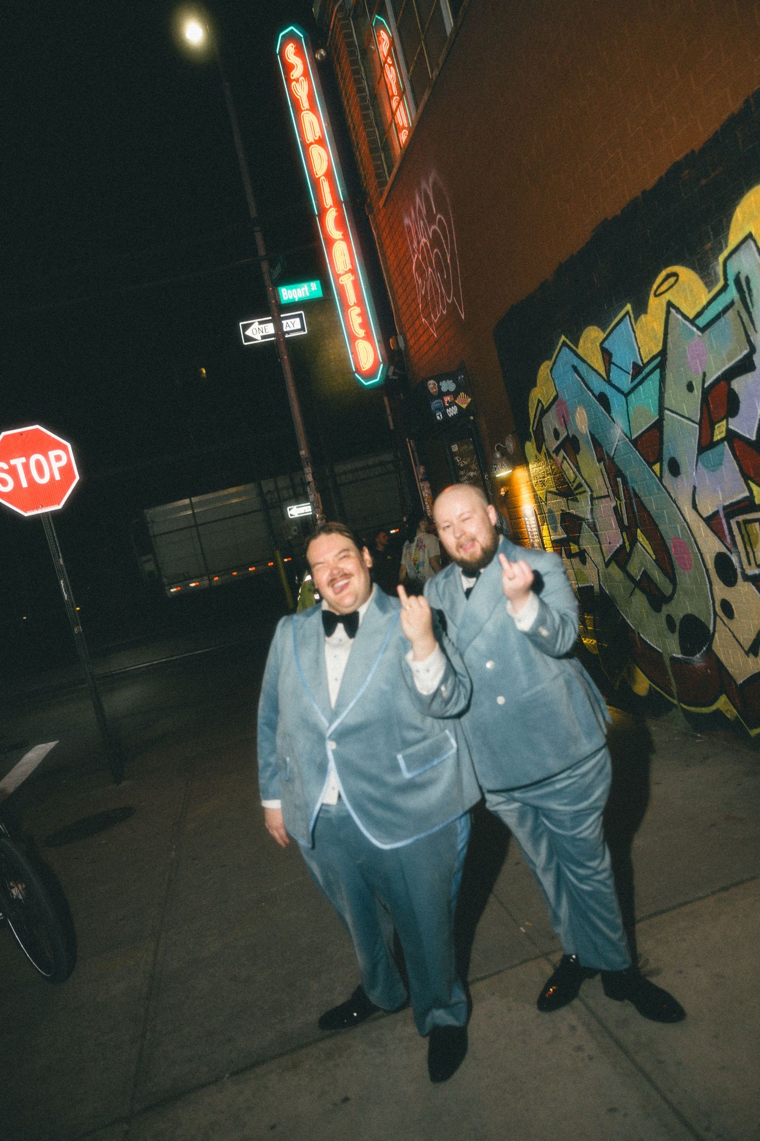 Two men in matching light blue velvet tuxedos pose on a city sidewalk at night near graffiti wall and neon signs.