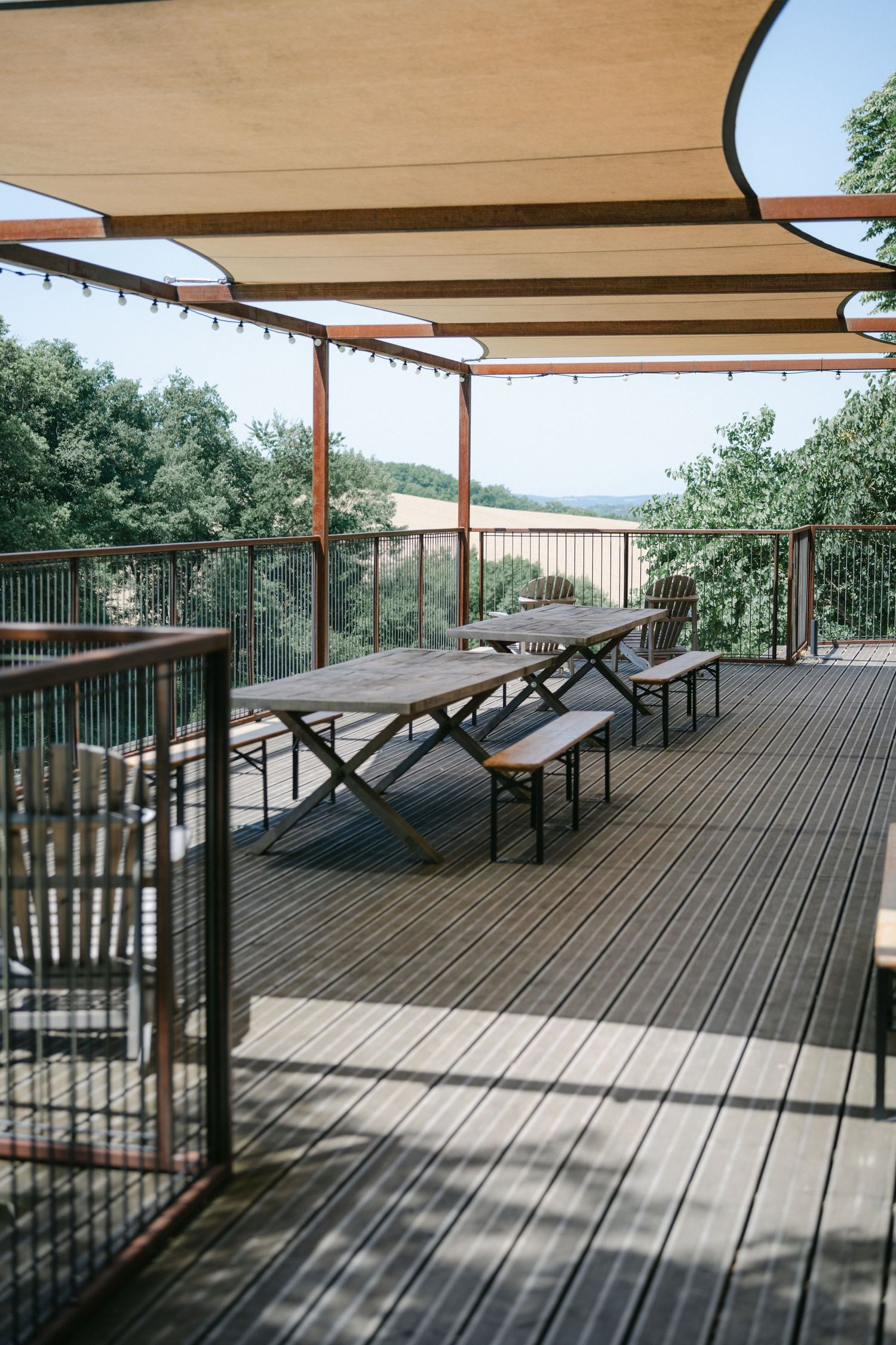 Outdoor wooden deck with picnic tables, shade sails, glass railing, and scenic green landscape view on a sunny day.