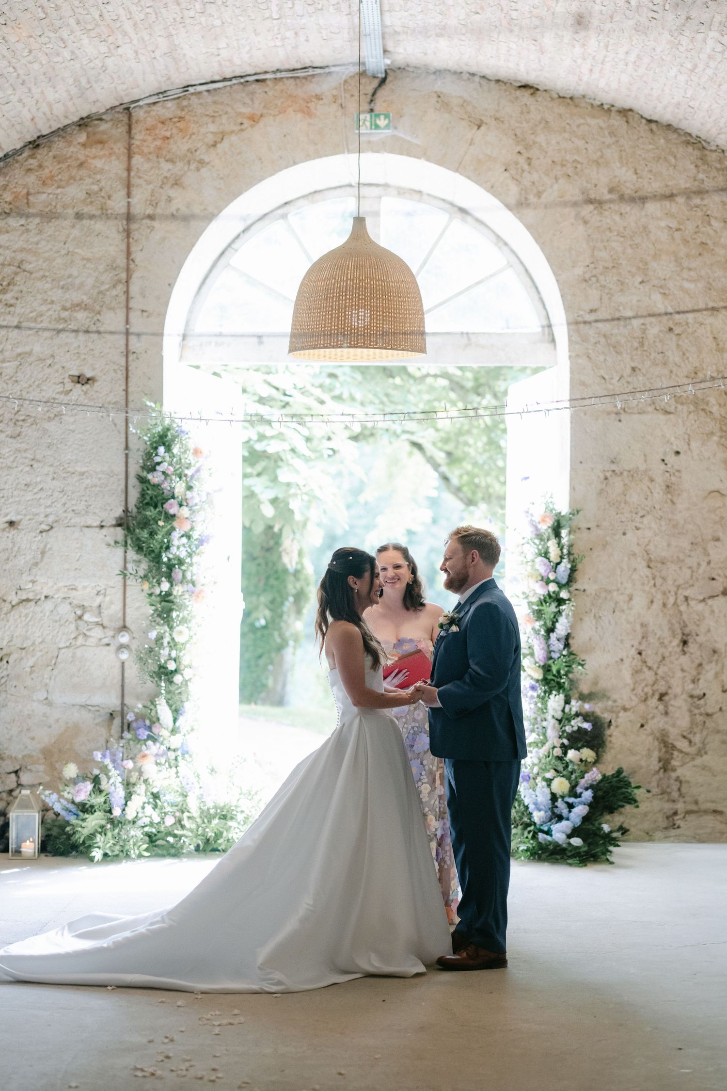 Bride and groom exchange vows before an officiant in a stone arch venue adorned with floral arrangements.