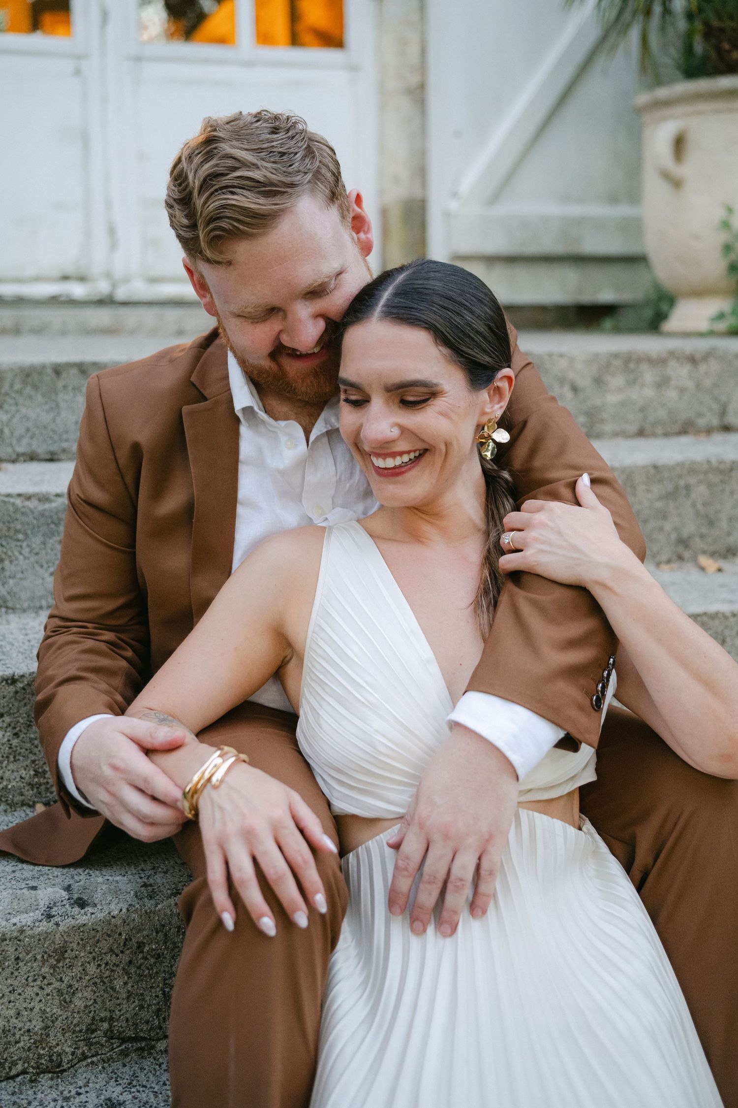 A smiling bride in a white dress sits on stone steps with a groom in a brown suit embracing her from behind.