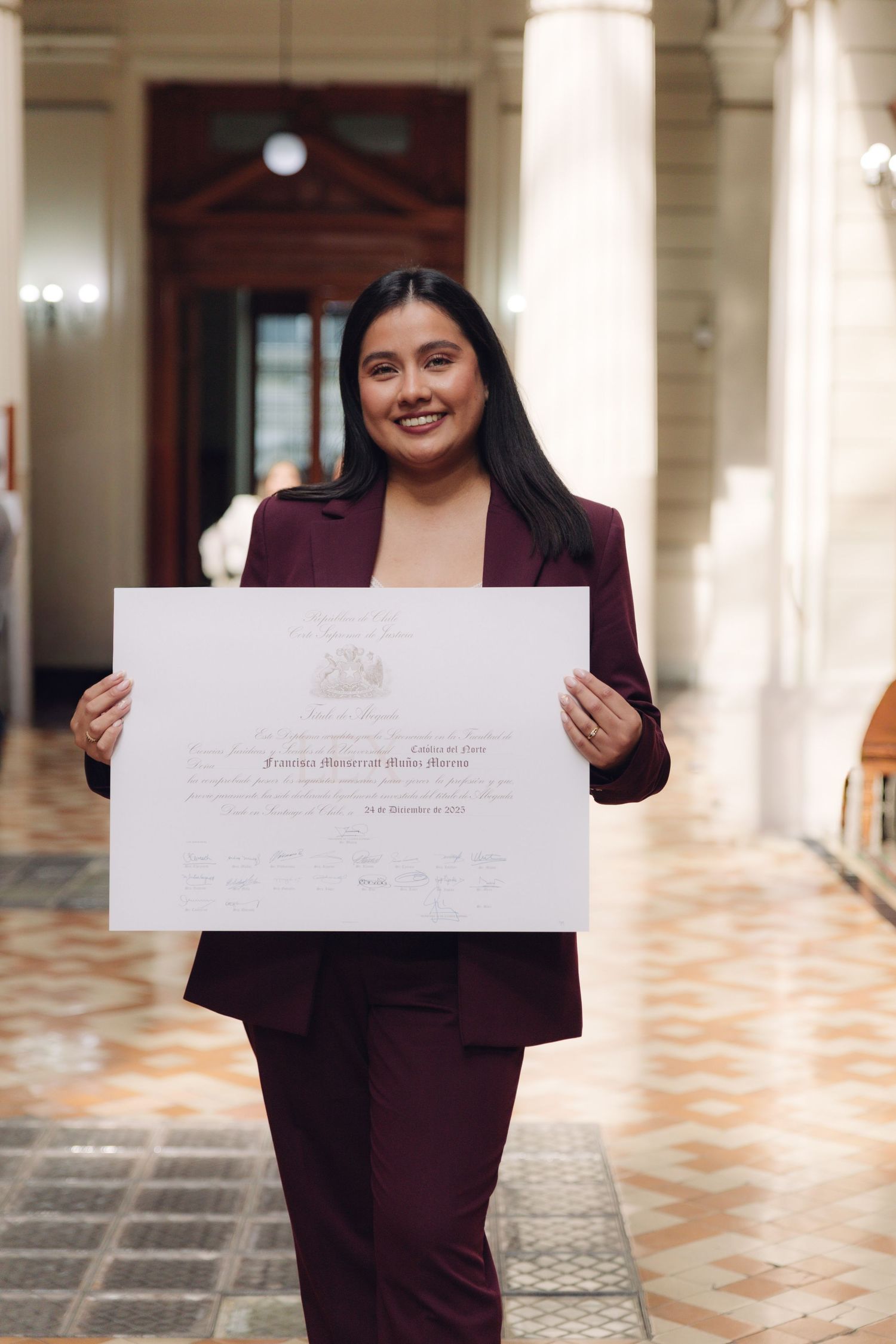 Smiling young woman in burgundy blazer holding a diploma certificate in a formal hall.