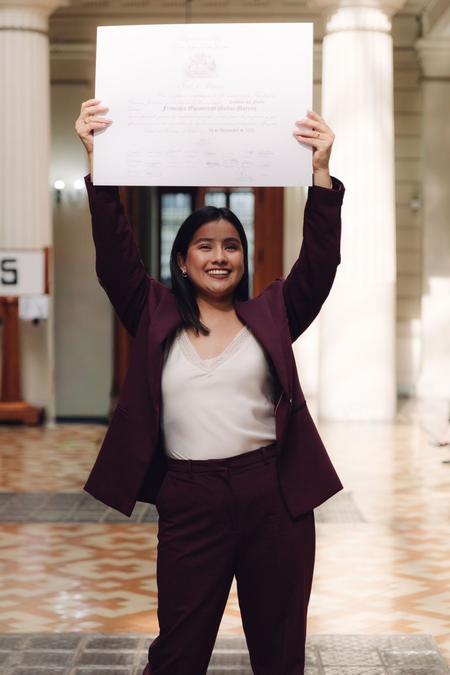 Joyful woman in maroon suit raising diploma certificate overhead in a grand hall.