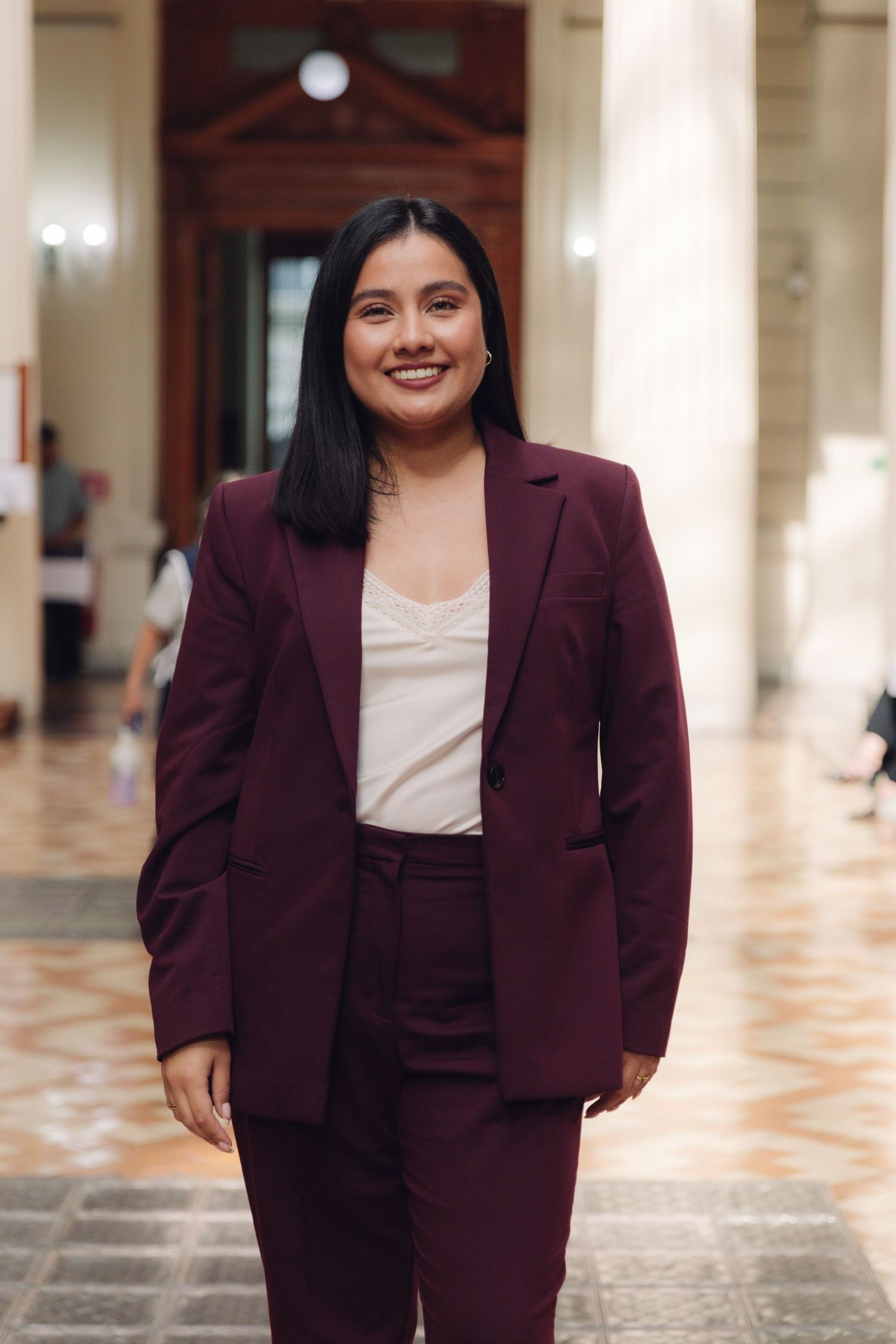 Confident young woman in burgundy suit smiling in an elegant marble-floored hall.