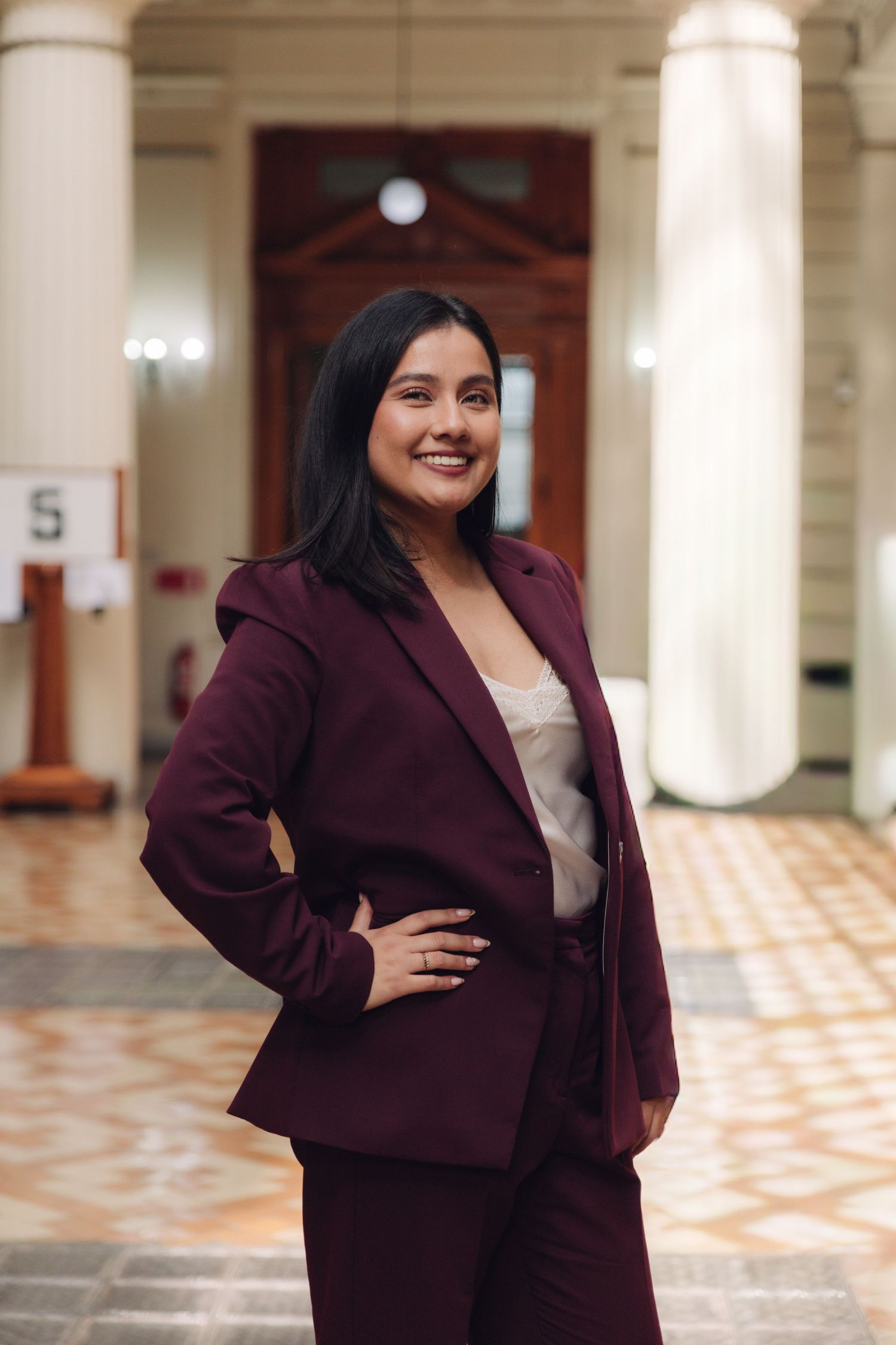 Professional woman in maroon blazer posing confidently in a classic interior hall.