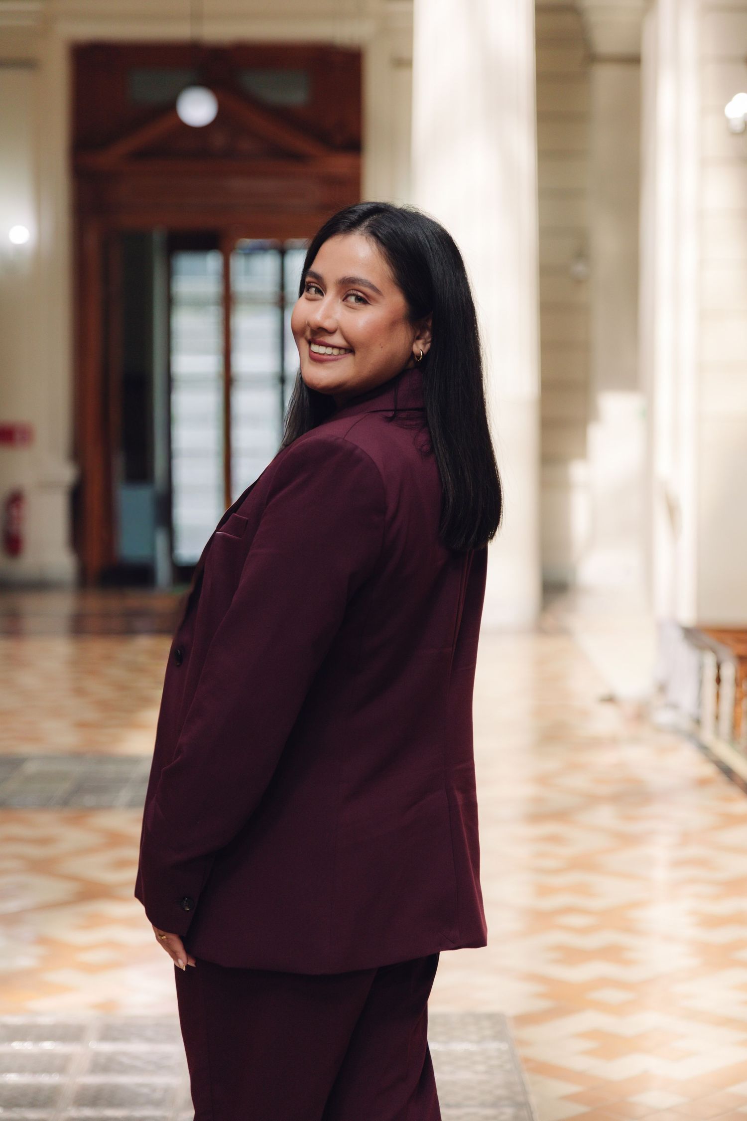 Smiling woman in burgundy suit looking back over shoulder in elegant building interior.