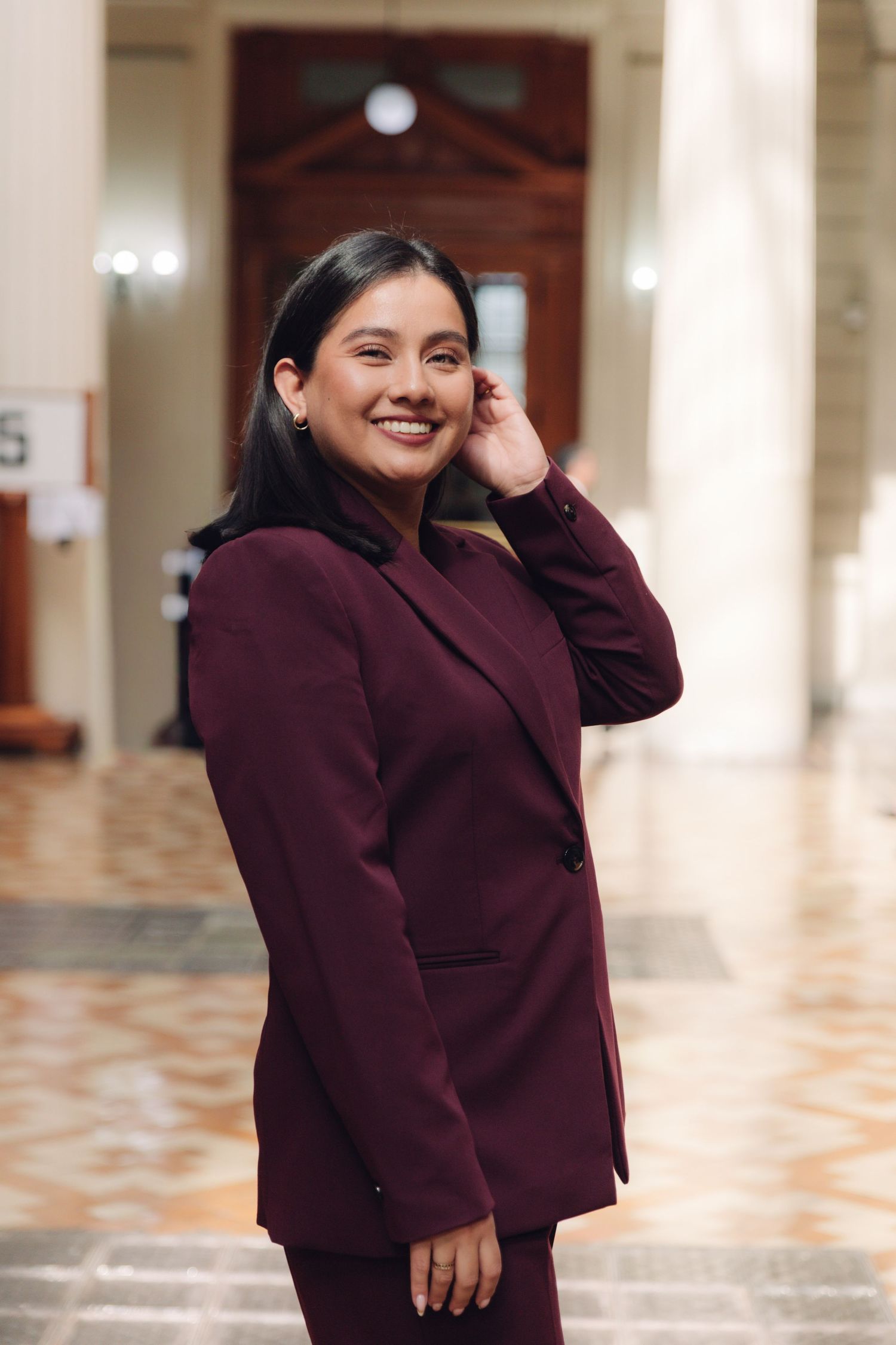 Young woman in burgundy suit smiling elegantly in a grand hallway.