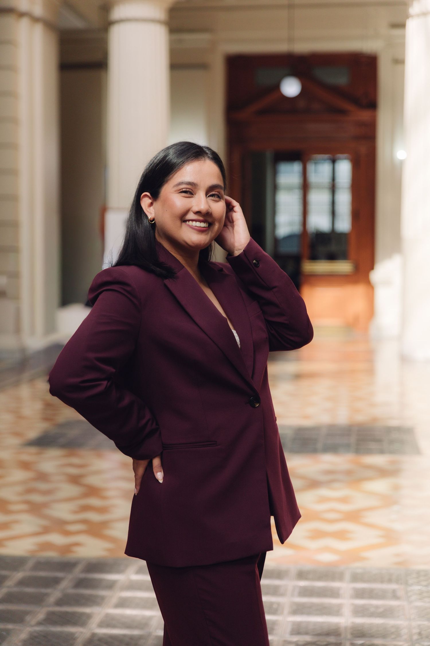 Professional woman in maroon suit posing confidently in ornate corridor.