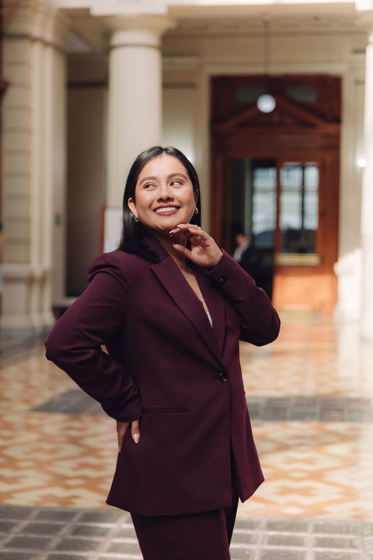 Smiling woman in dark red blazer standing in elegant building interior.