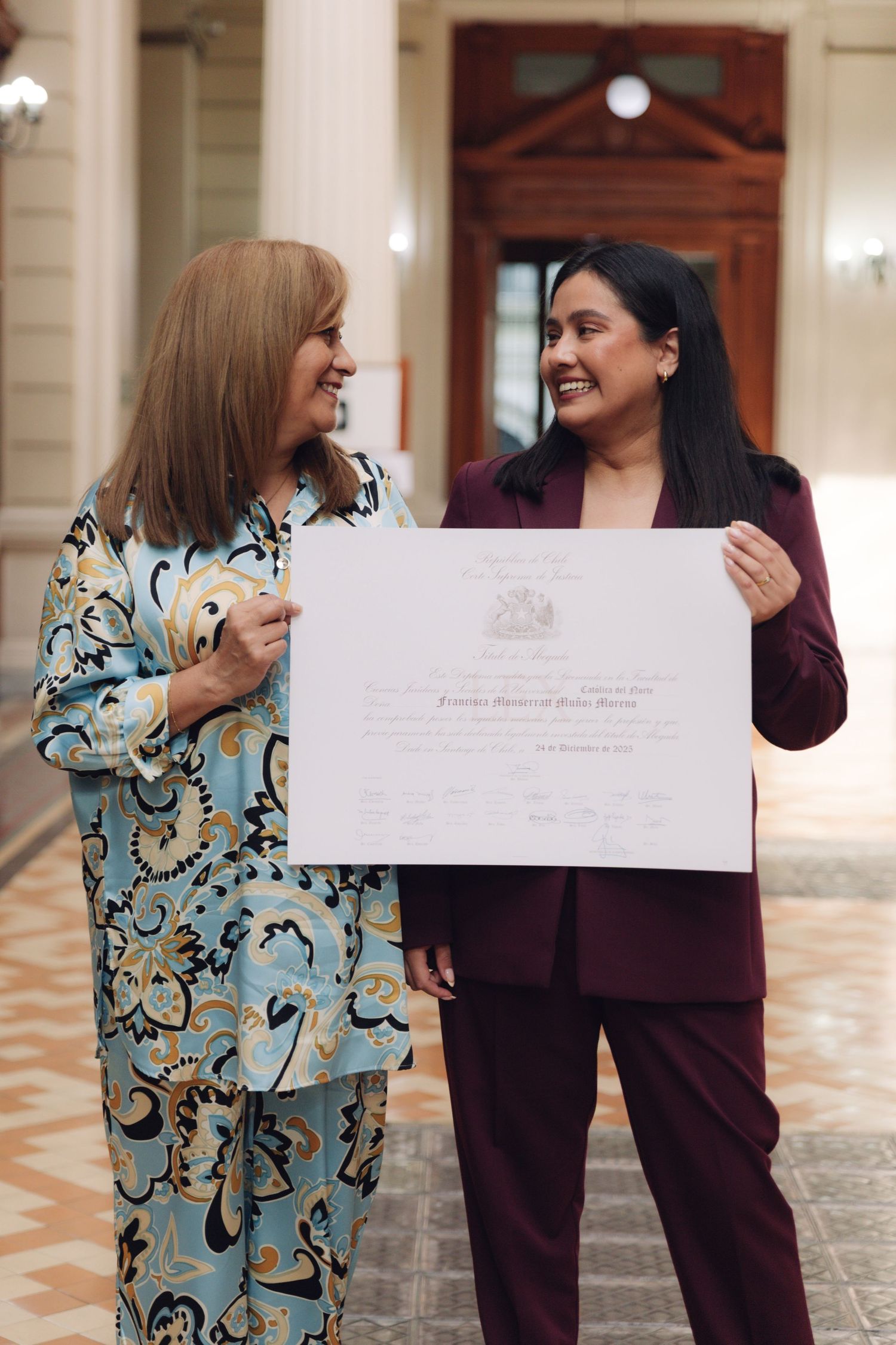 Two women proudly holding a formal certificate in a classic hall.