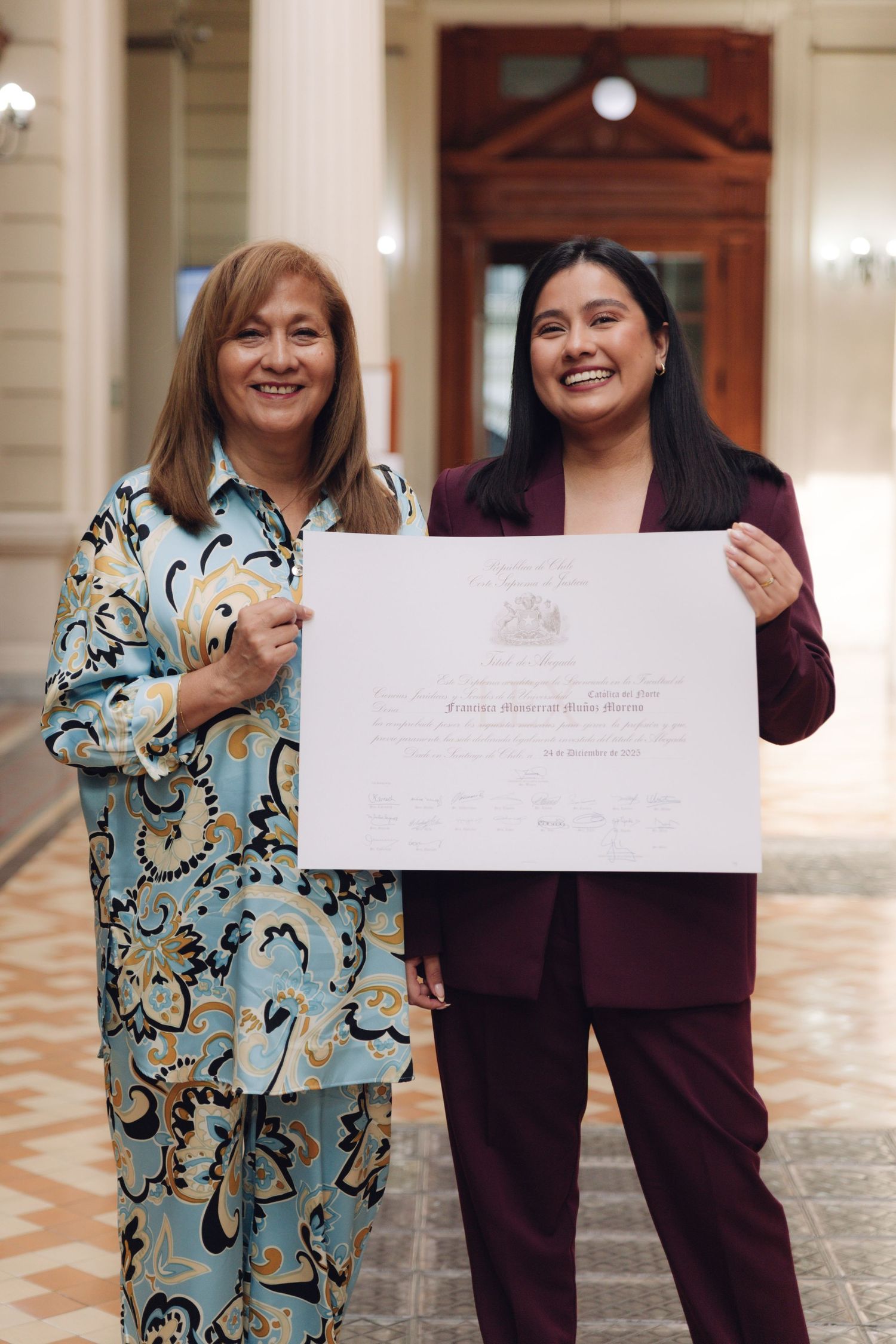 Two women smiling and displaying an official diploma in grand hallway.