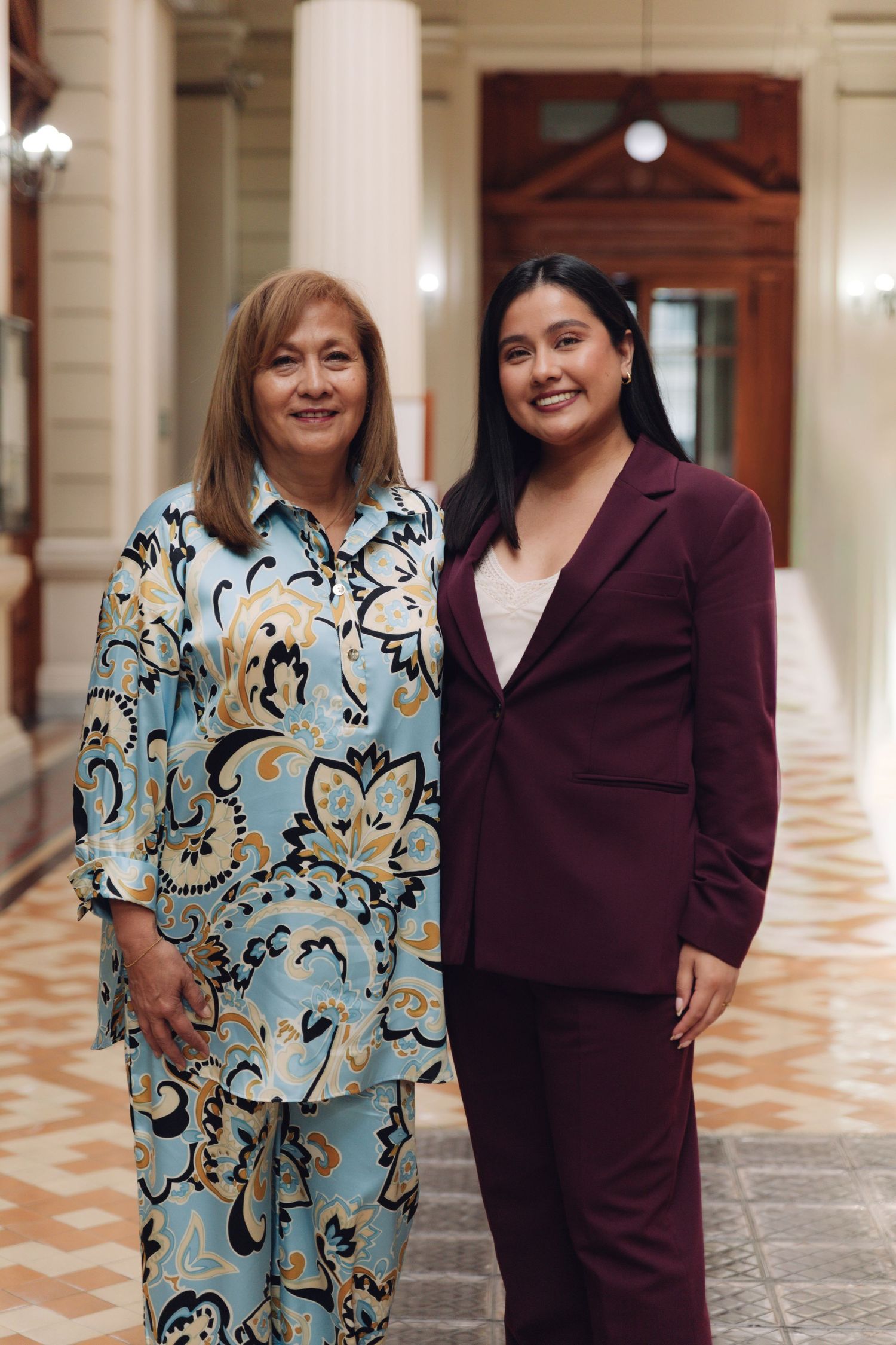 Two women posing together smiling in an elegant columned corridor.
