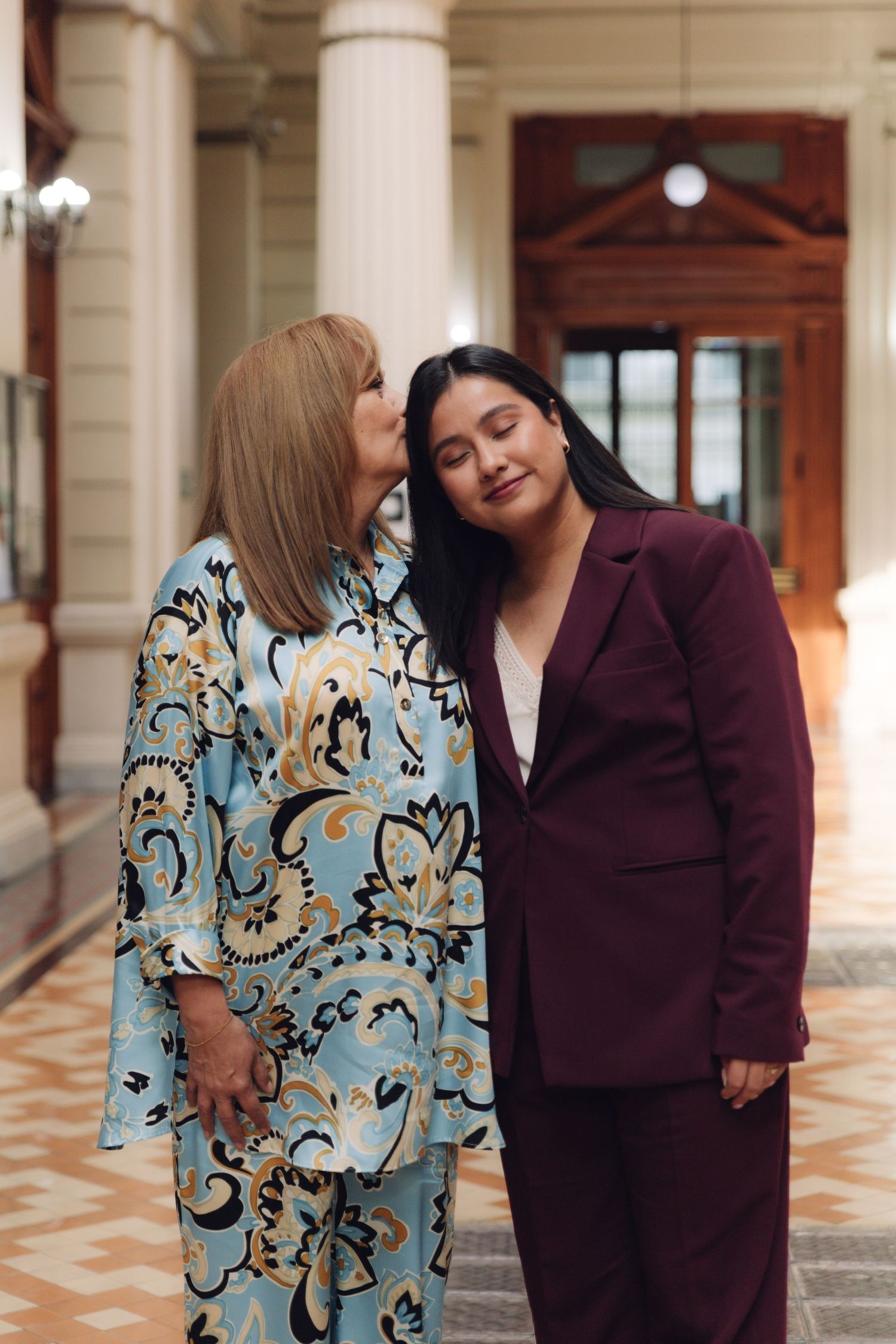 Older woman affectionately kissing younger woman in burgundy suit indoors.