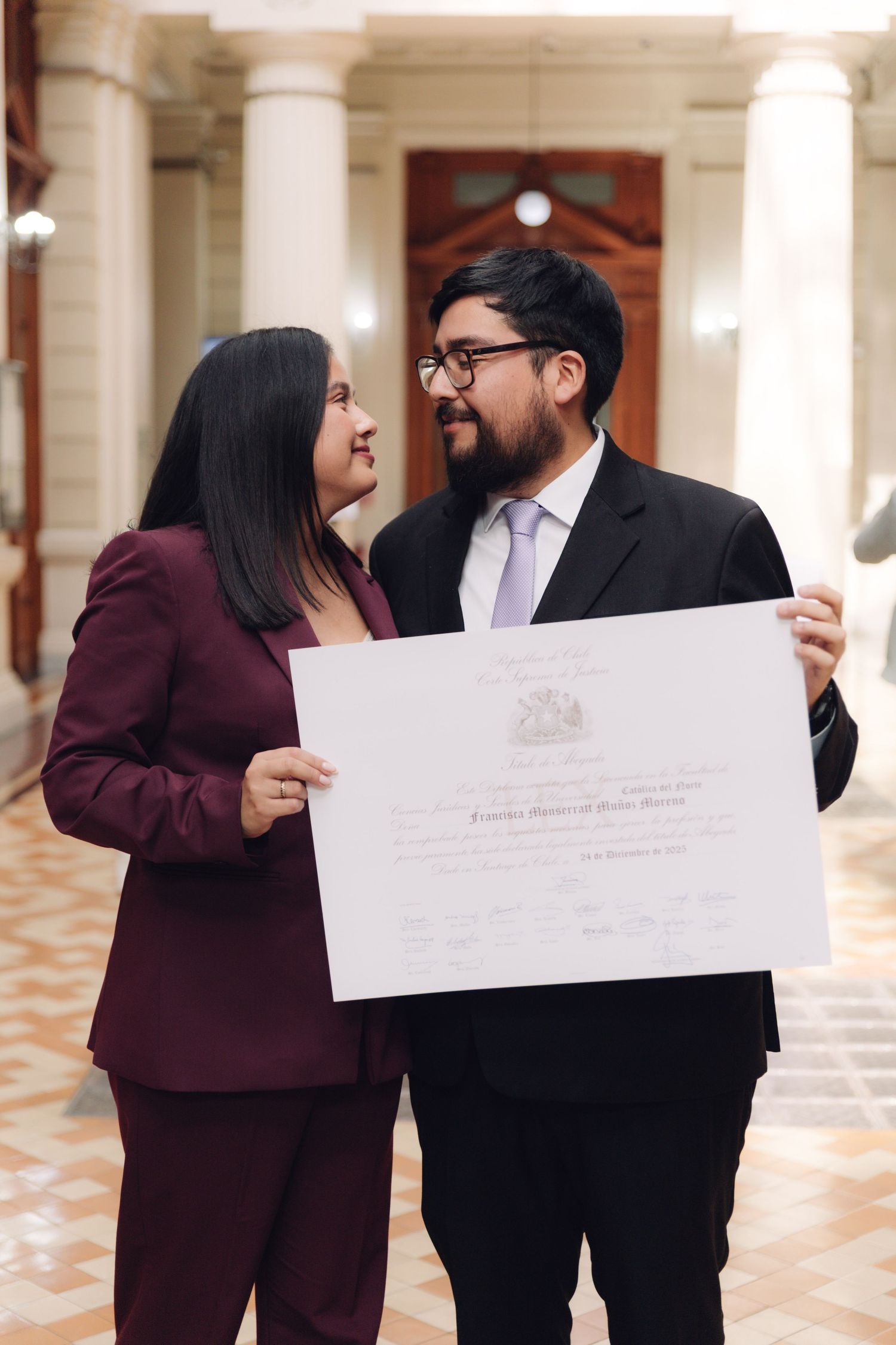 Couple smiling at each other while holding official certificate in hallway.