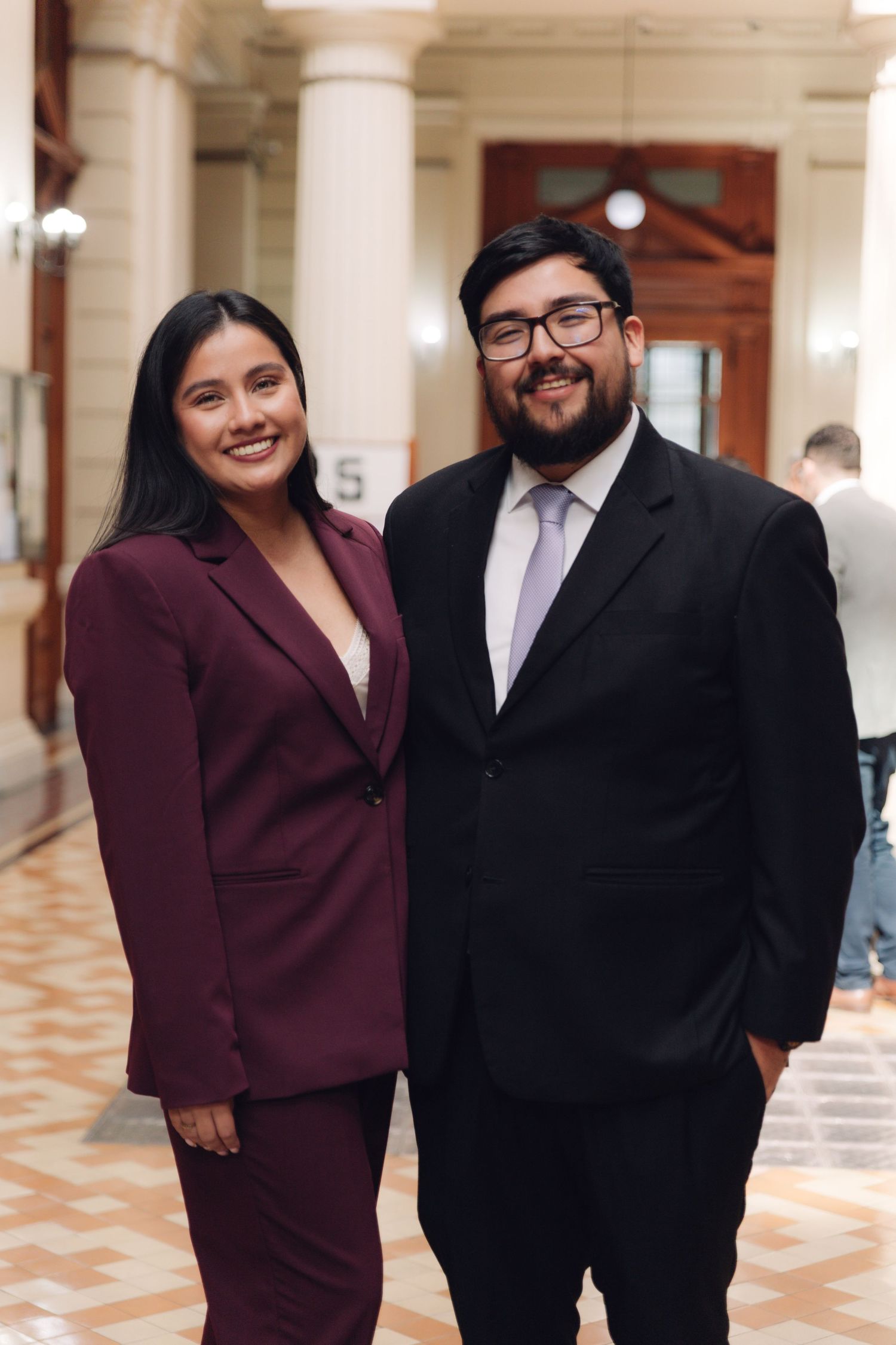 Young woman in burgundy suit and man in black suit smiling together in elegant hallway.