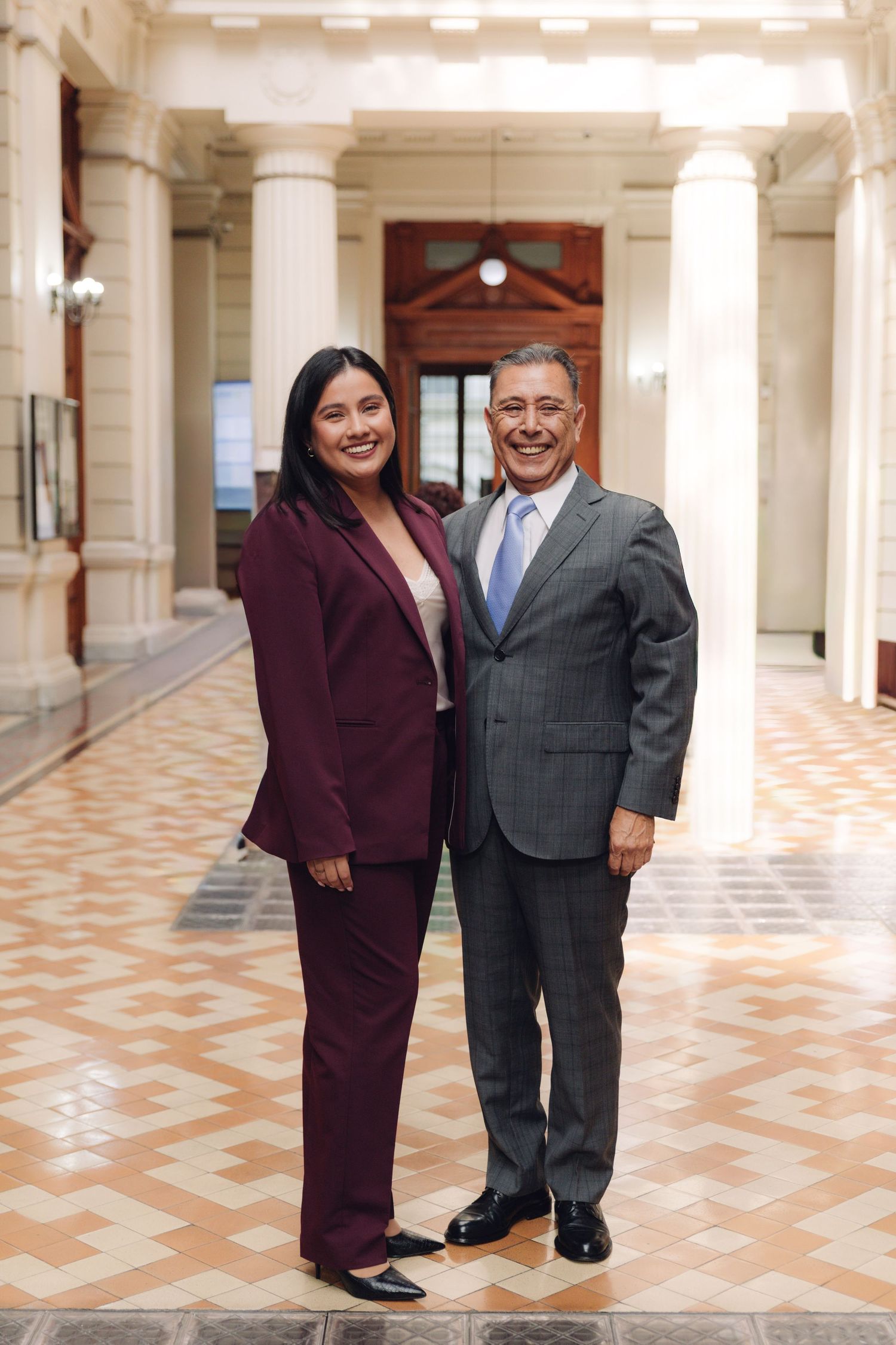 Woman in burgundy suit and older man in gray suit posing together in formal building corridor.