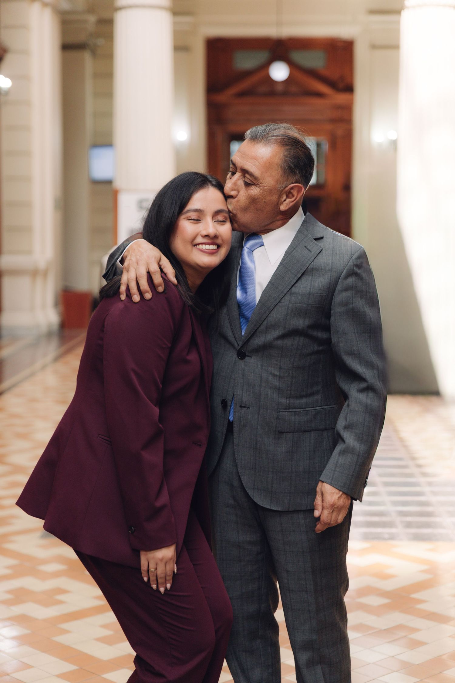Older man in gray suit kissing smiling woman in burgundy suit on cheek in hallway.