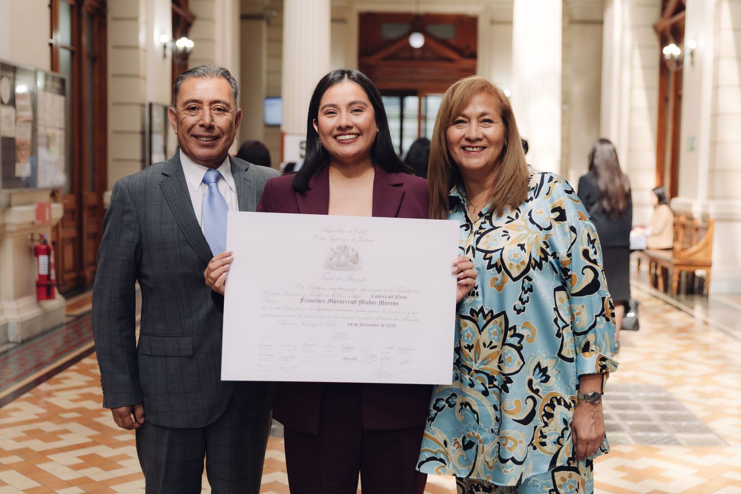 Woman in burgundy suit holding certificate flanked by older man and woman in floral top.