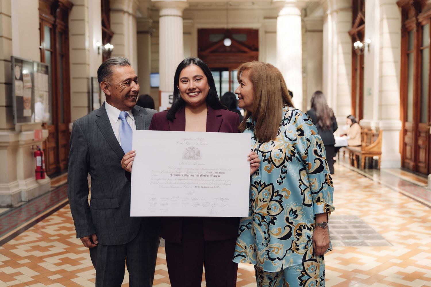 Three people smiling together while examining an official certificate in a formal building.