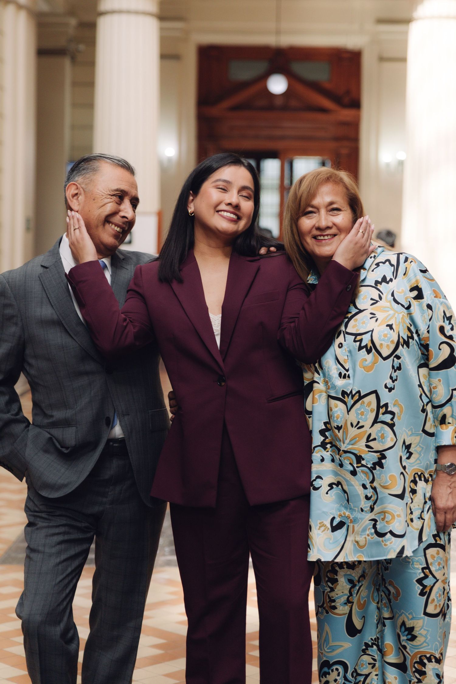 Woman in burgundy suit playfully touching cheeks of older man and woman in floral blouse.