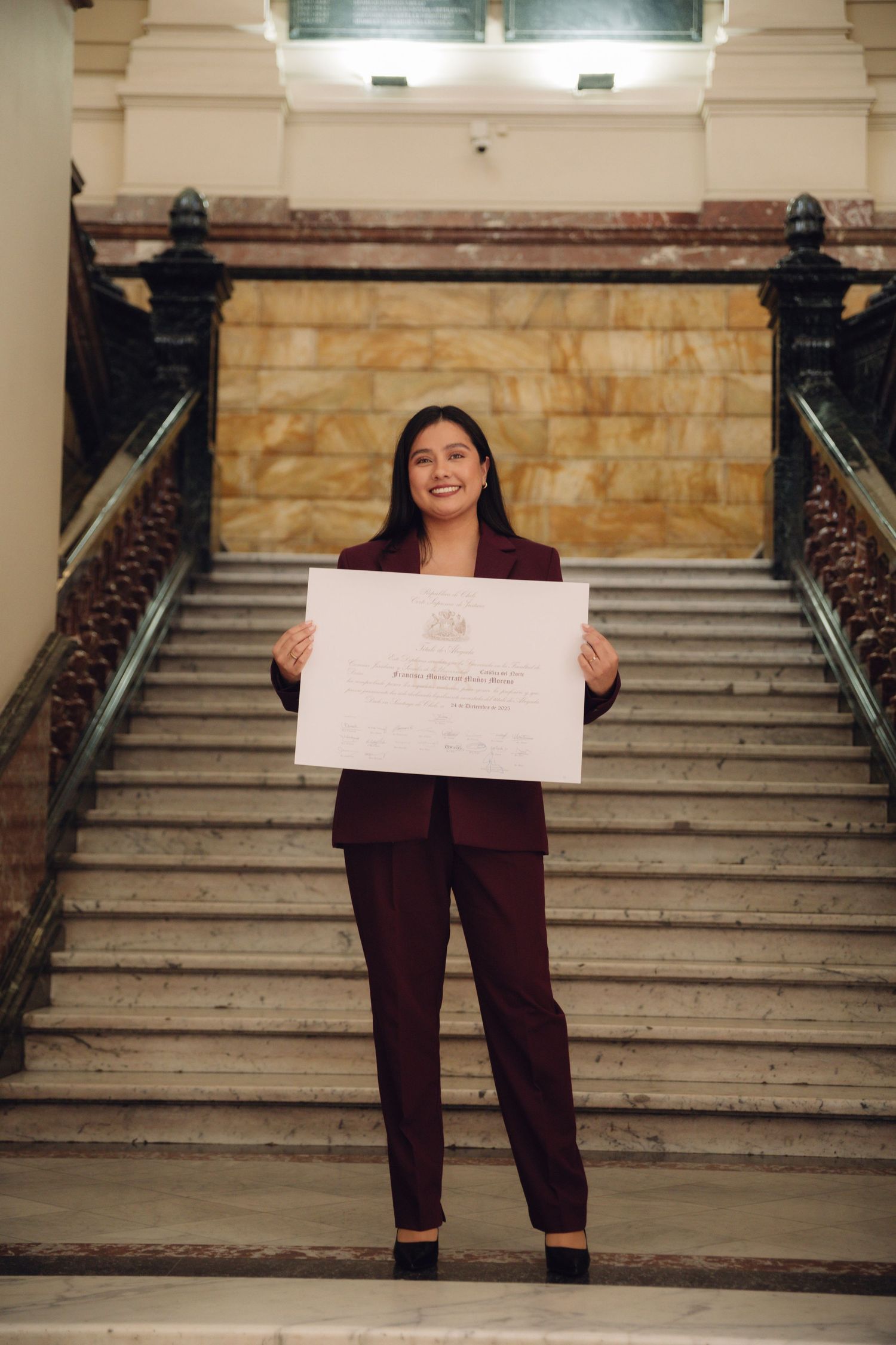 Happy woman in burgundy suit proudly displaying official certificate on elegant staircase.