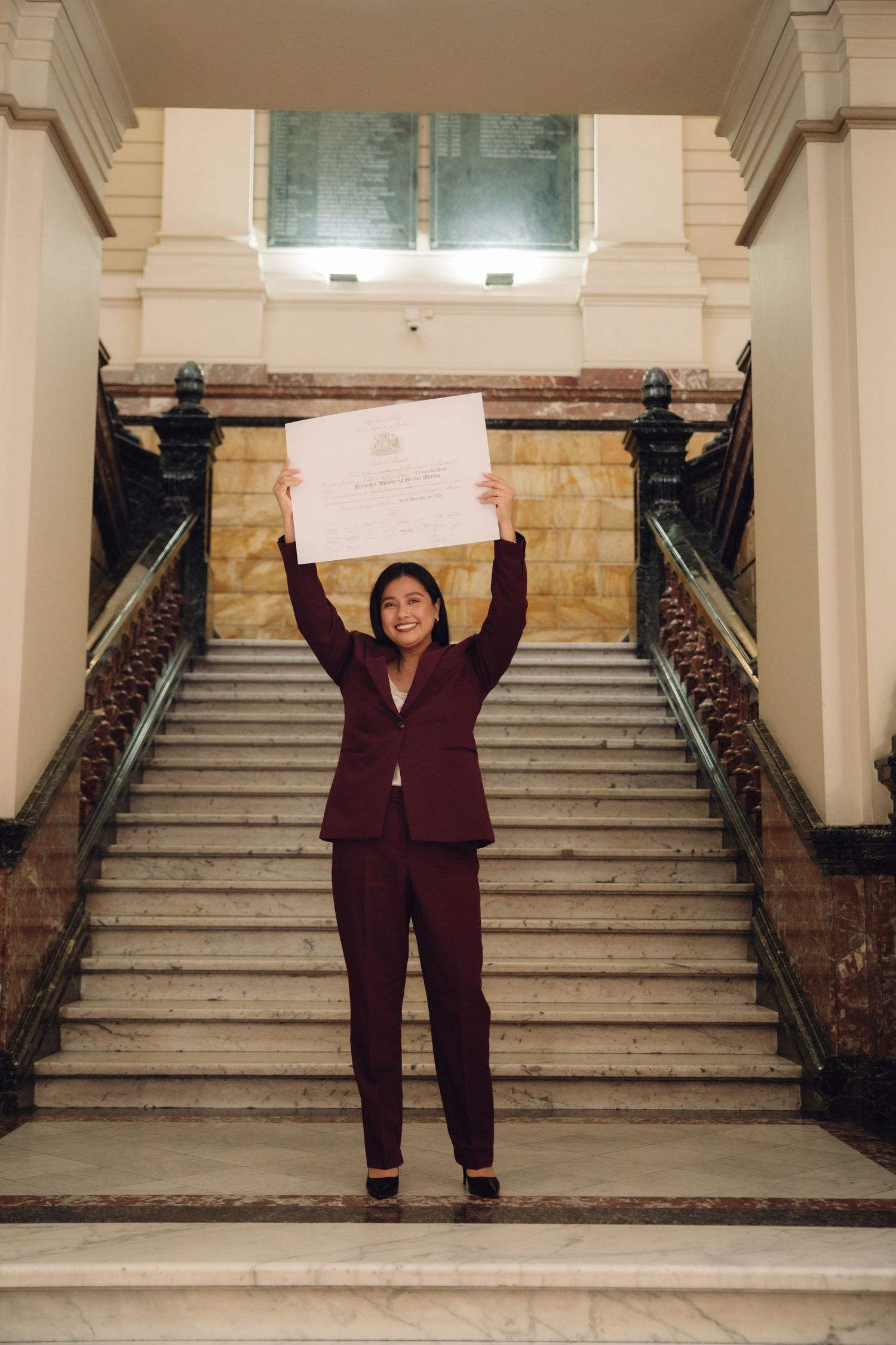 Joyful woman in burgundy suit raising official certificate triumphantly on grand staircase.