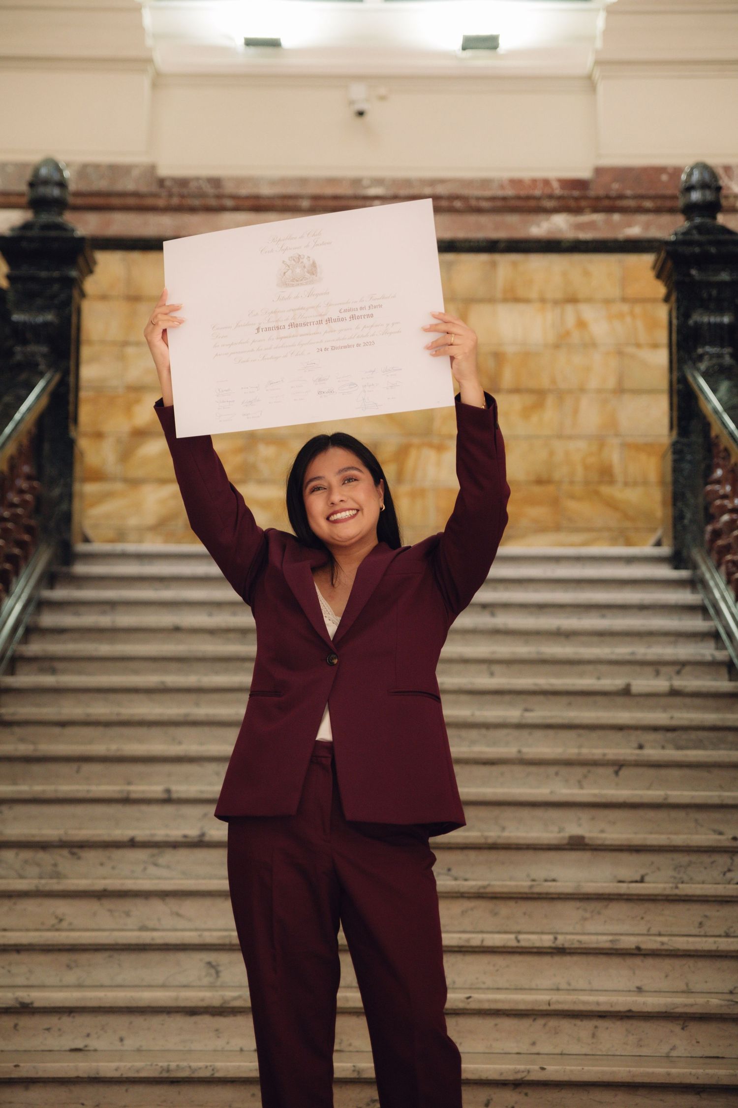 Young woman in burgundy suit joyfully raises diploma overhead on grand staircase.