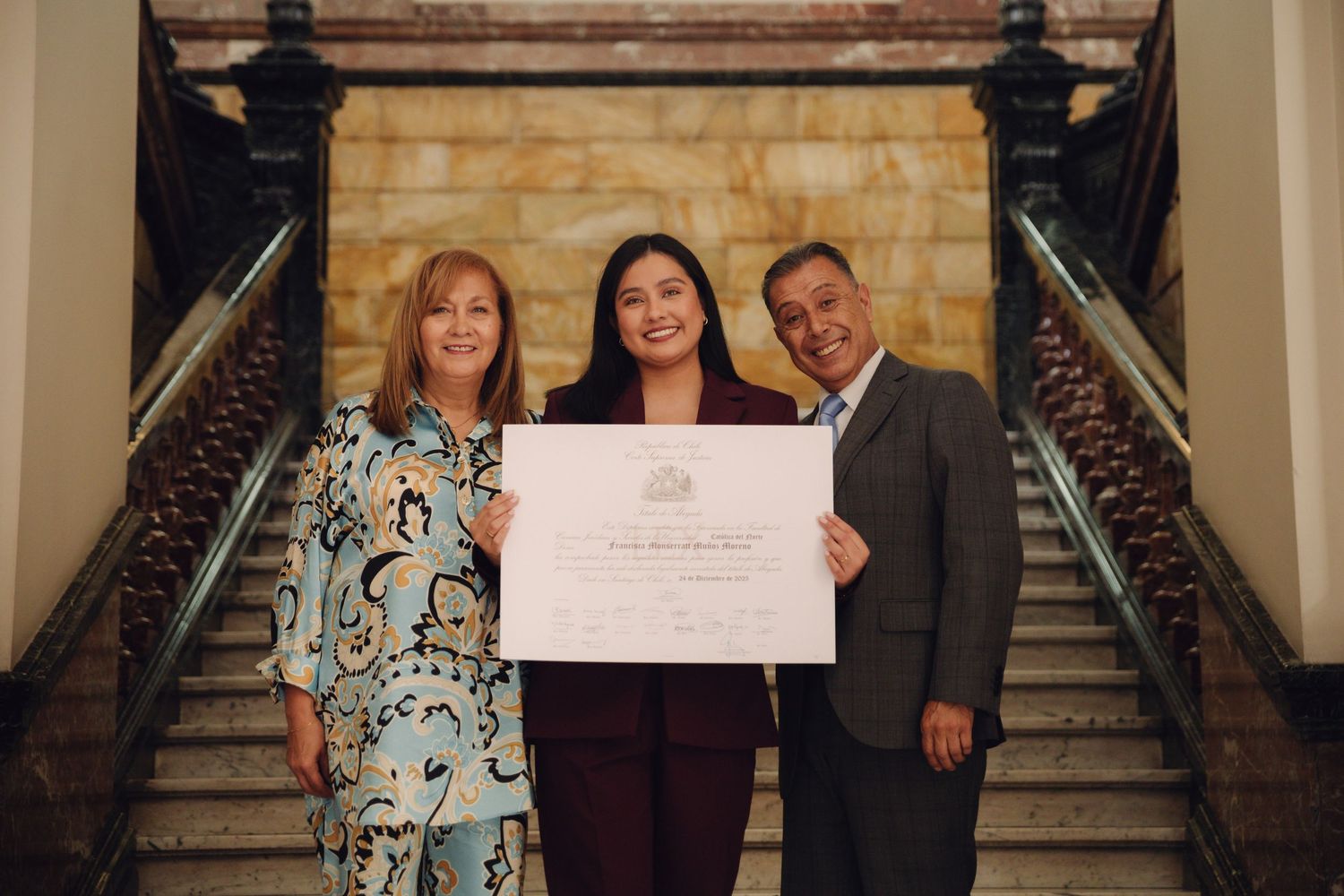 Graduate in burgundy suit proudly holds diploma flanked by proud parents on staircase.