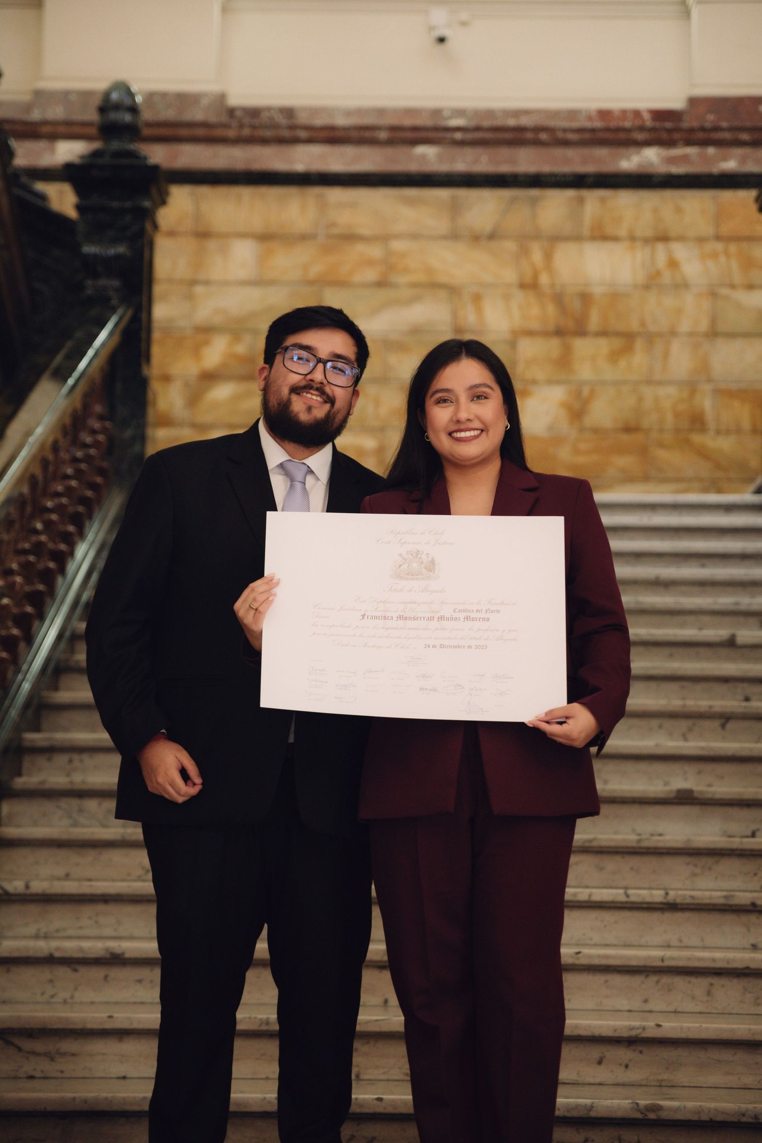 Smiling graduate in burgundy suit holds diploma certificate with suited companion on stairs.