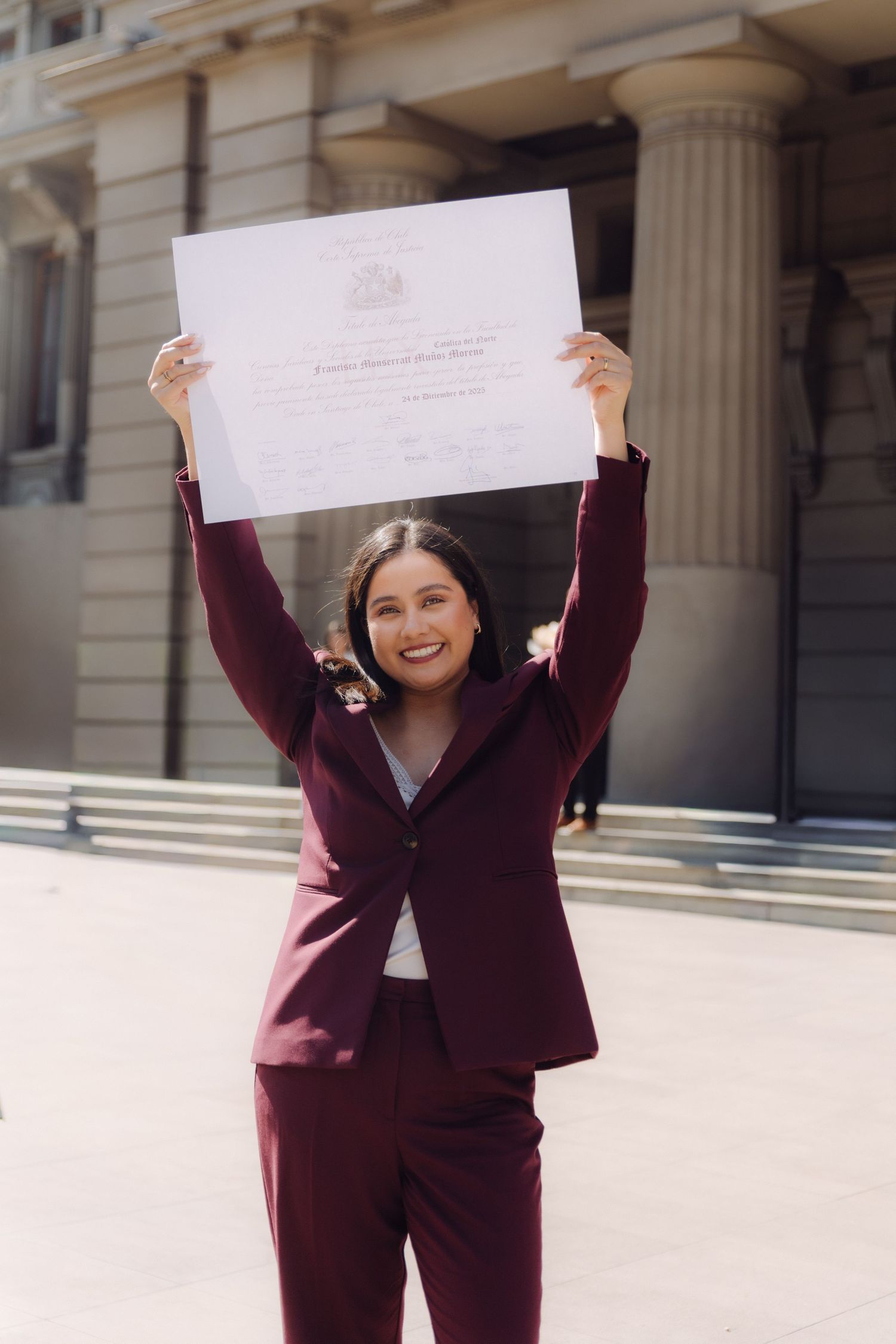 Jubilant woman in maroon suit raises graduation diploma outdoors near classical building.