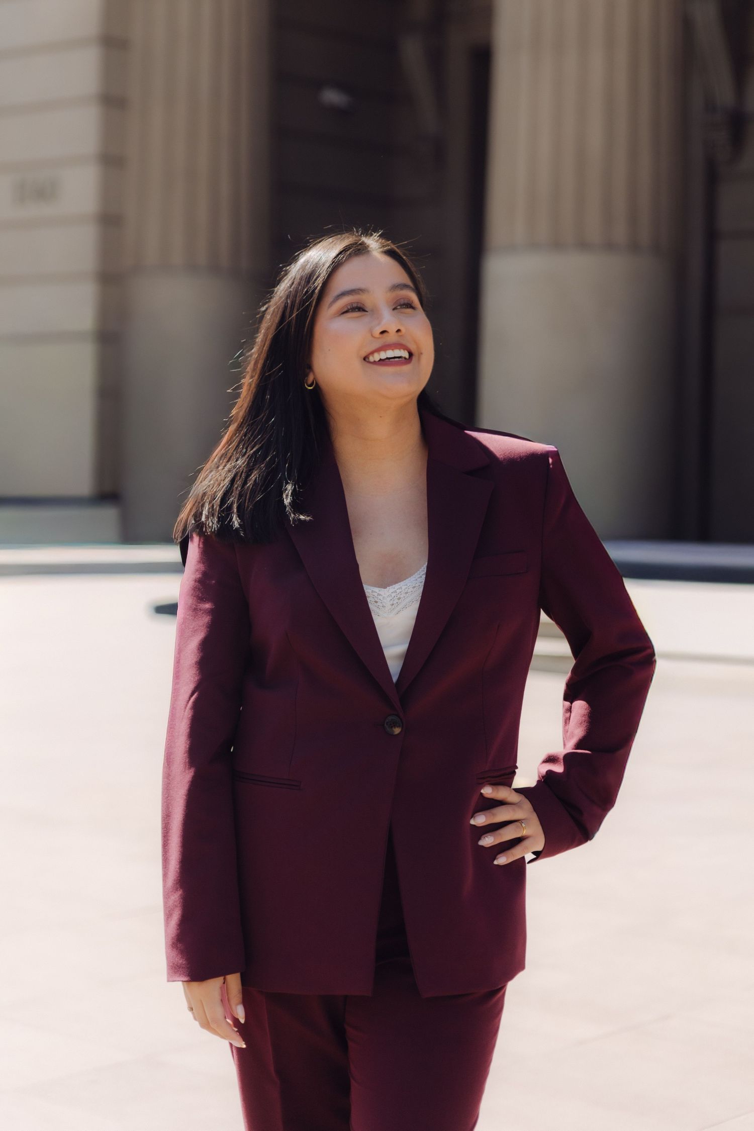 Confident young woman in burgundy blazer smiles outdoors near classical stone columns.