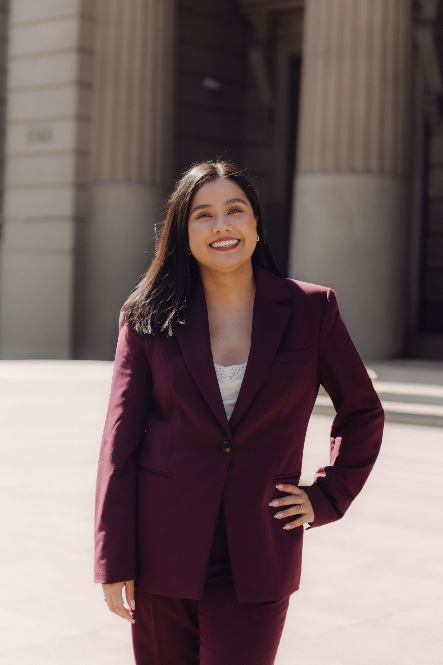 Smiling professional woman in maroon suit poses with hand on hip before building.