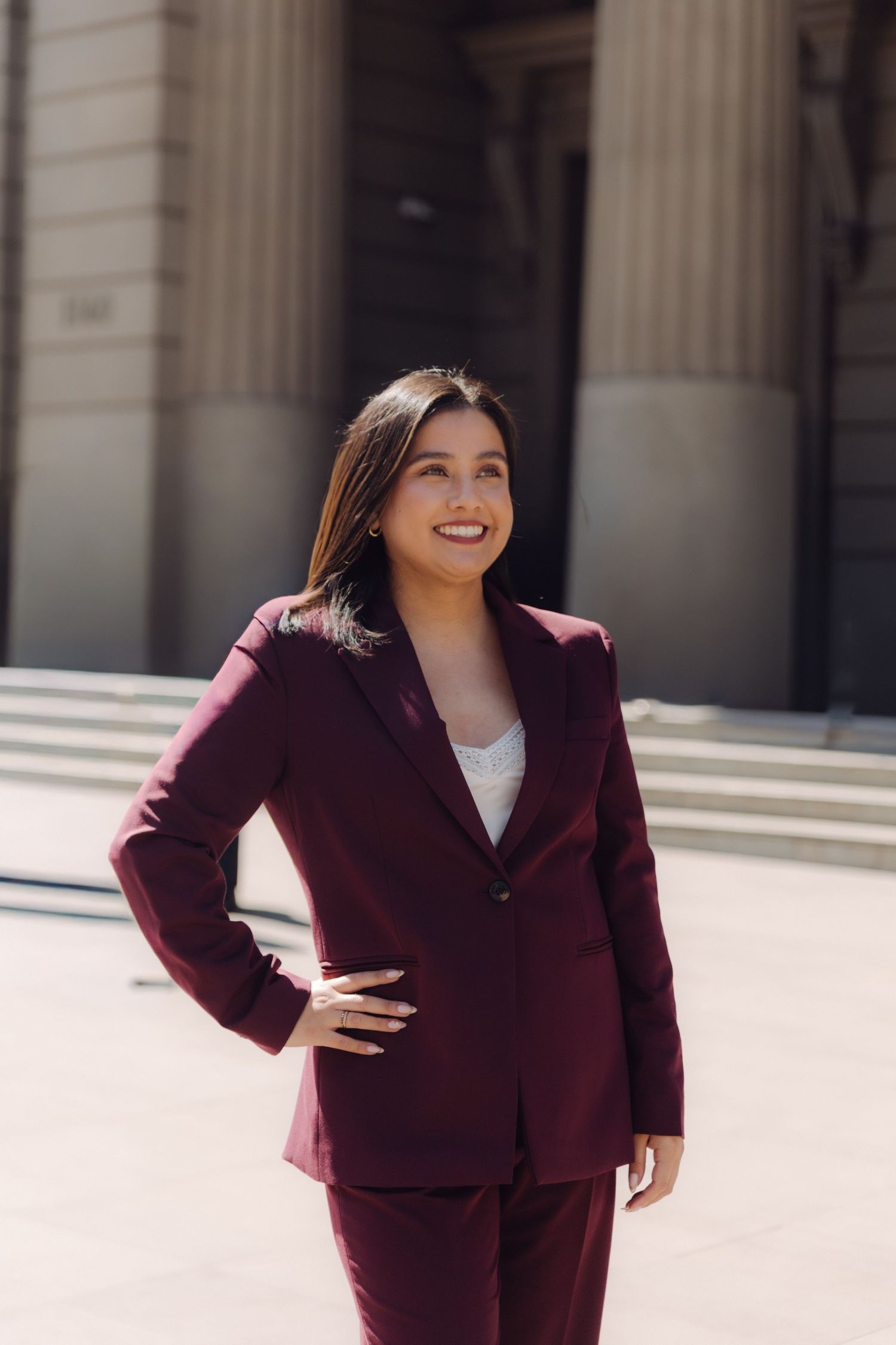 Elegant young woman in burgundy suit smiles confidently outside classical architecture.