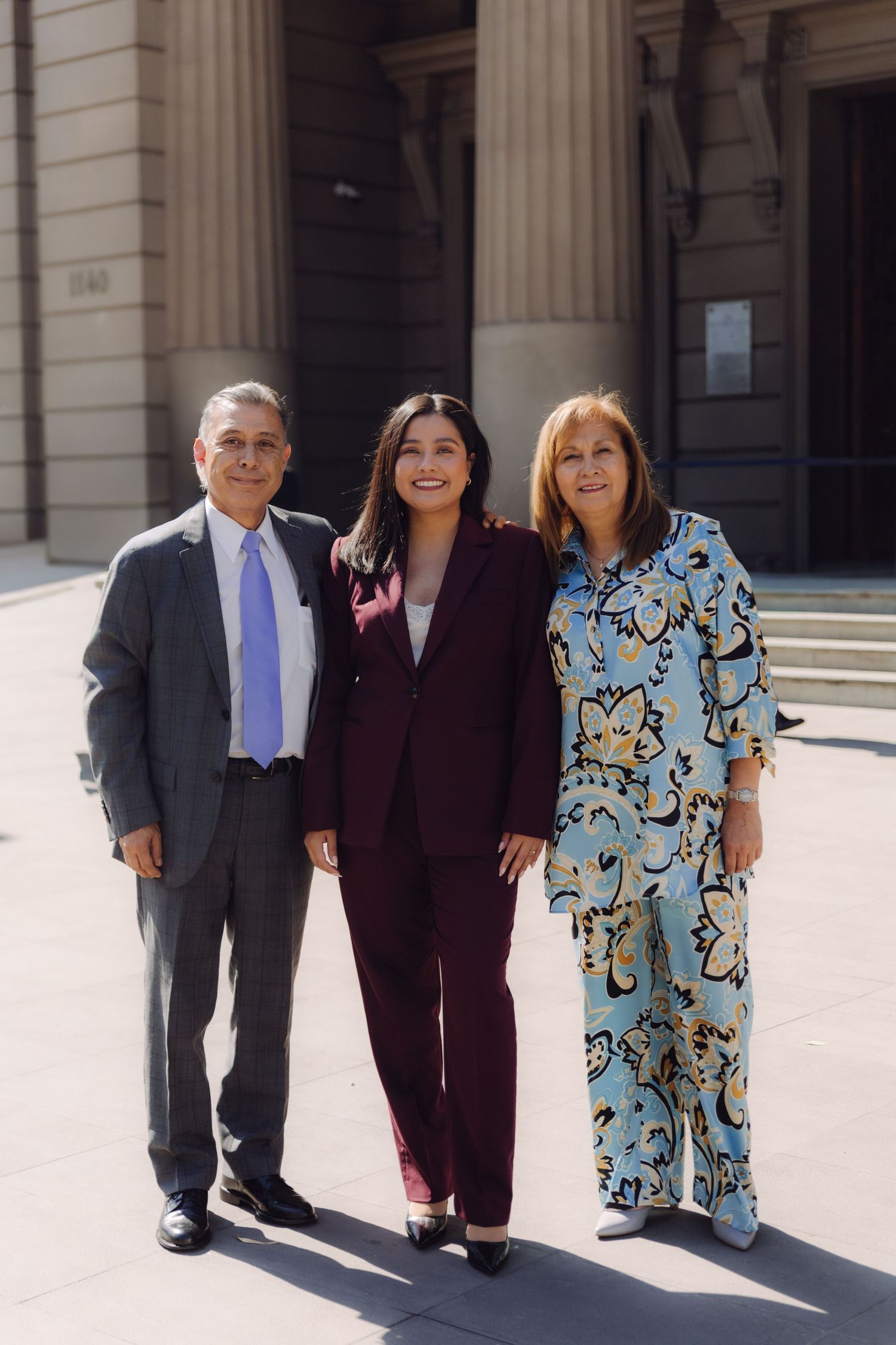 Graduate in burgundy suit poses proudly with parents outside neoclassical building.