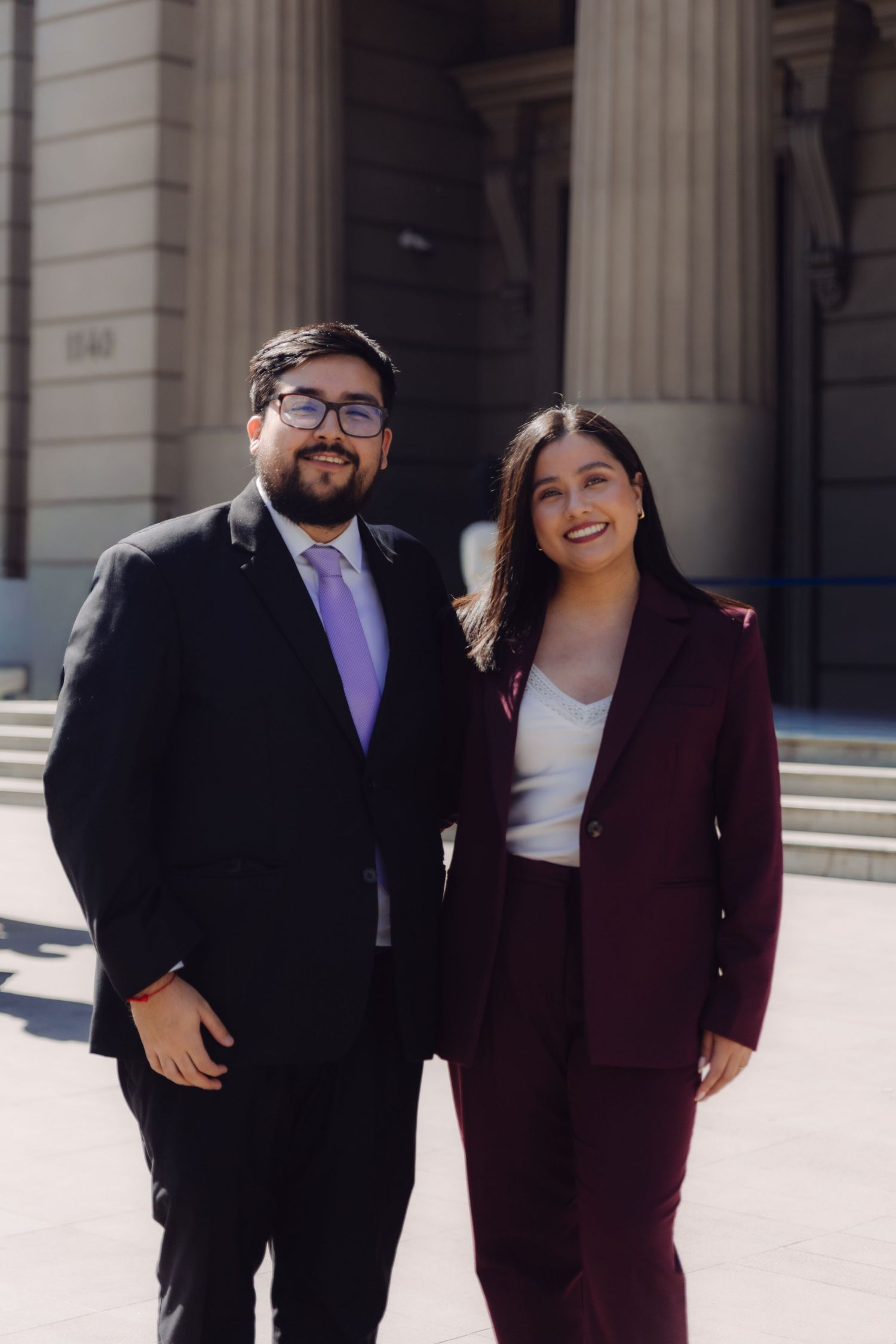Smiling graduate in burgundy suit stands with bespectacled companion outside stone building.