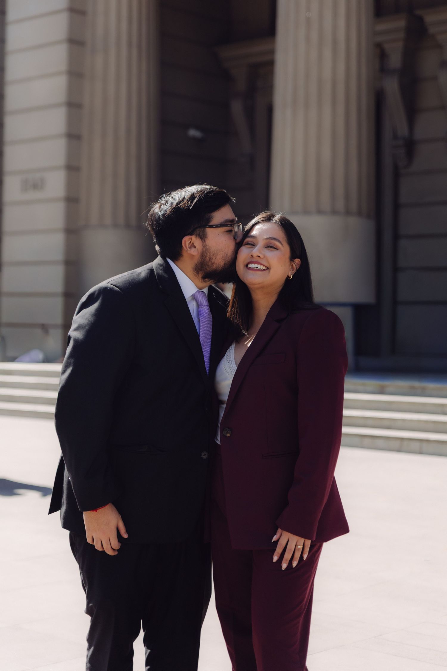 Couple in formal attire sharing a kiss outside a grand building, man in black suit and woman in burgundy pantsuit.