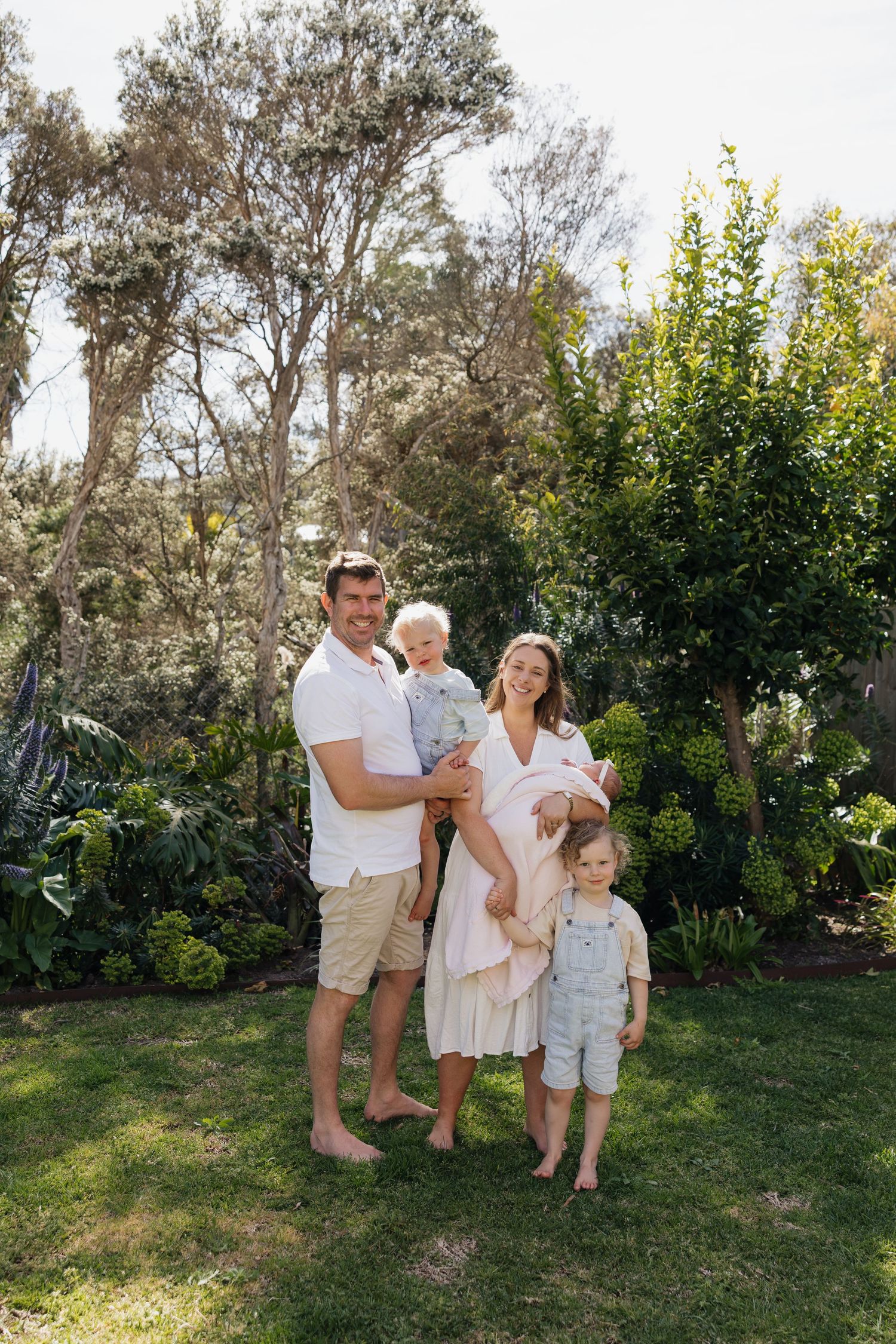 Family of five with newborn baby posing barefoot on green lawn surrounded by lush garden trees in natural light.