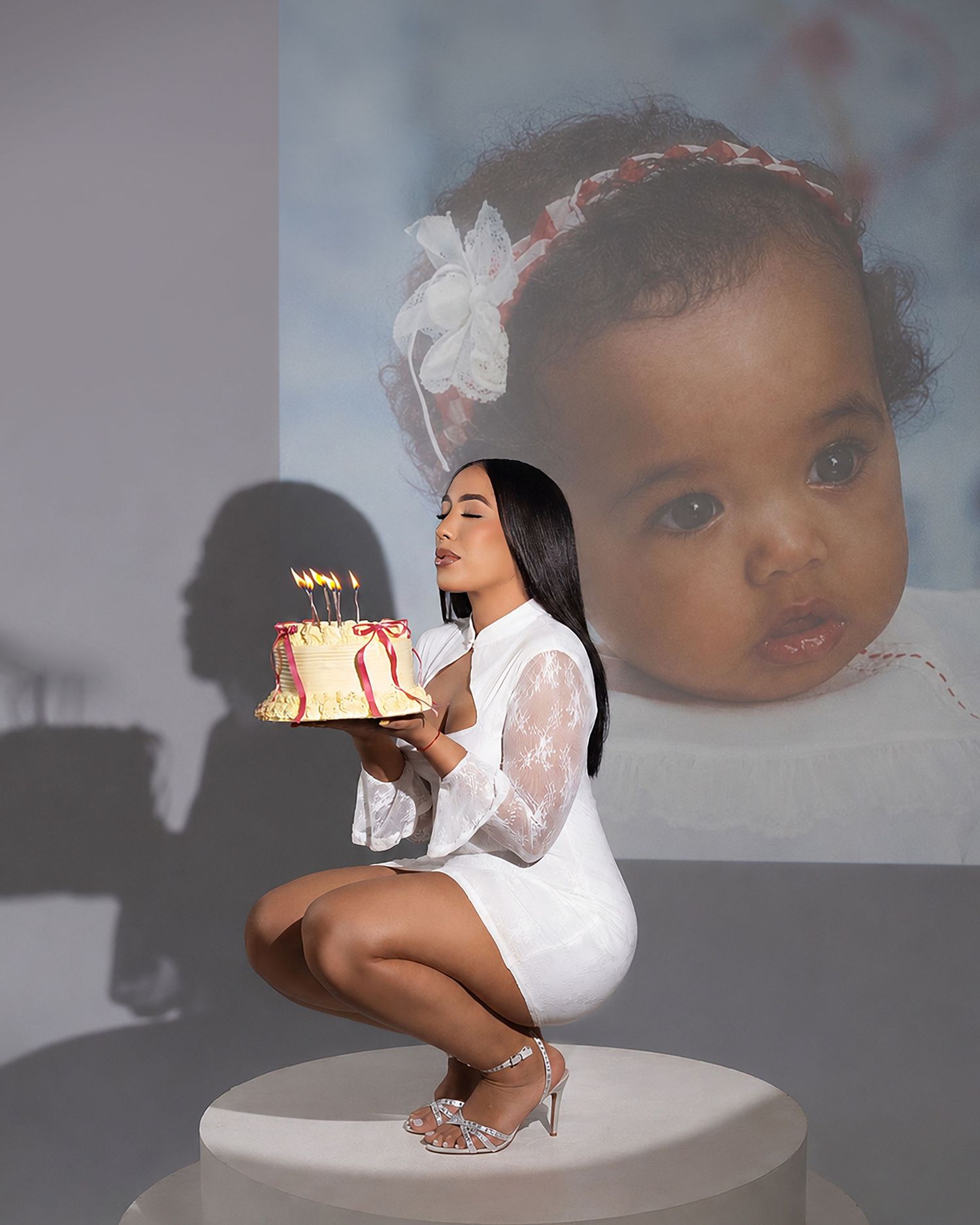 Woman in white dress blowing birthday cake candles, with a large baby photo projected on the wall behind her.