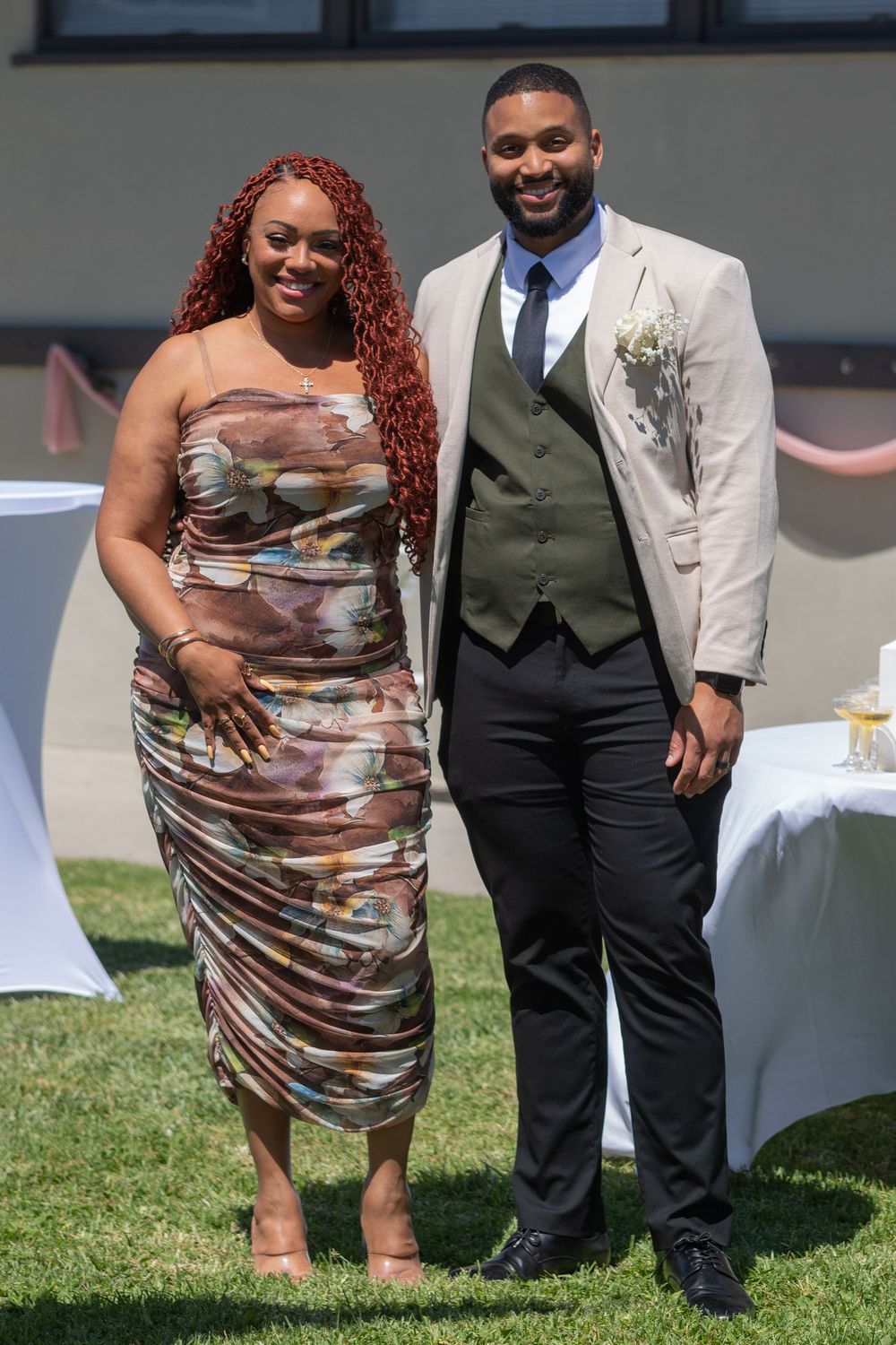 A couple posing outdoors at a wedding reception, woman in floral dress with red hair, man in tan blazer and vest.