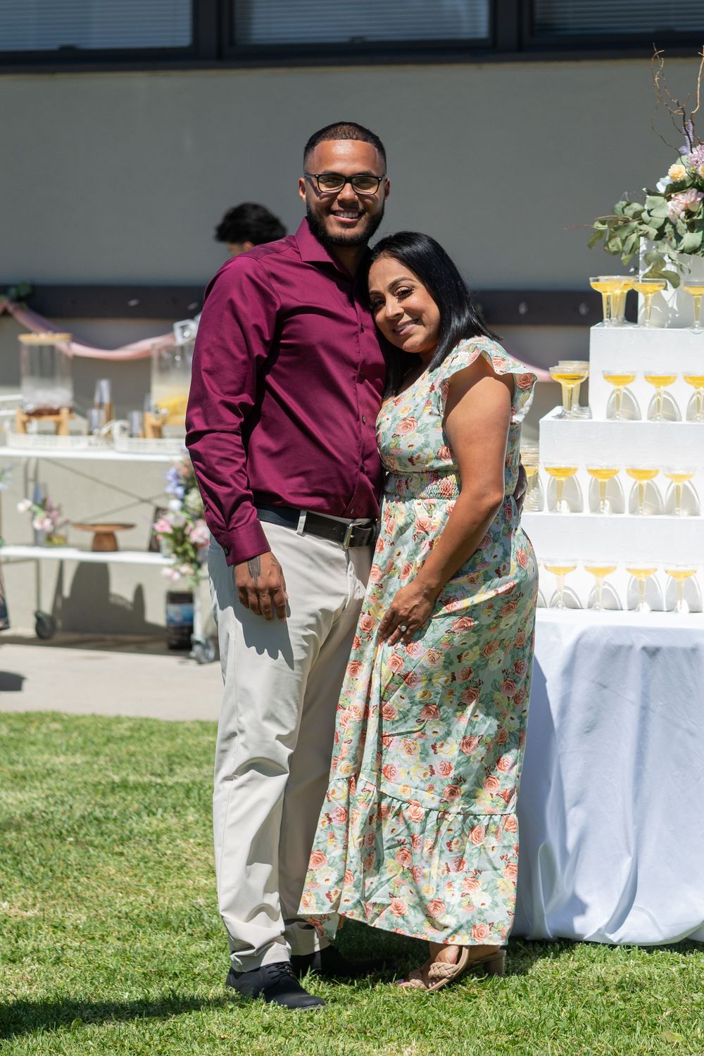 Couple smiling together at an outdoor event, man in maroon shirt and woman in floral dress near a decorated table.