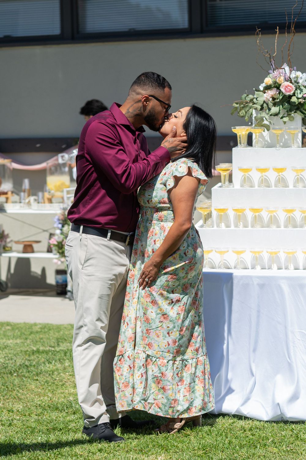 Couple sharing a romantic kiss at an outdoor celebration near a decorated dessert table with floral arrangements.