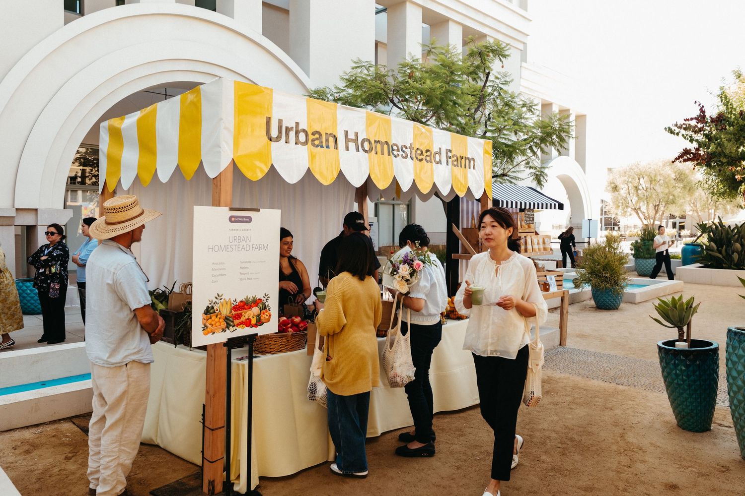 Visitors browse the Urban Homeland Farms market stall with yellow striped awning at an outdoor event.